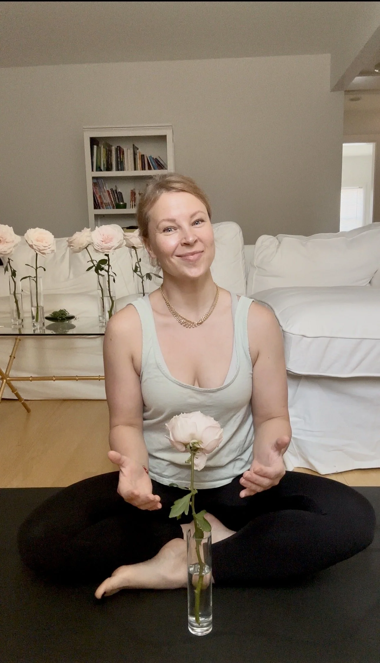 Woman sitting cross-legged holding a pink rose in front of her, with a tall glass vase containing the same pink rose on a black surface in front. She is smiling and giving a thumbs-up with both hands. She is in a living room with white couch, bookshelves, and minimal decor.