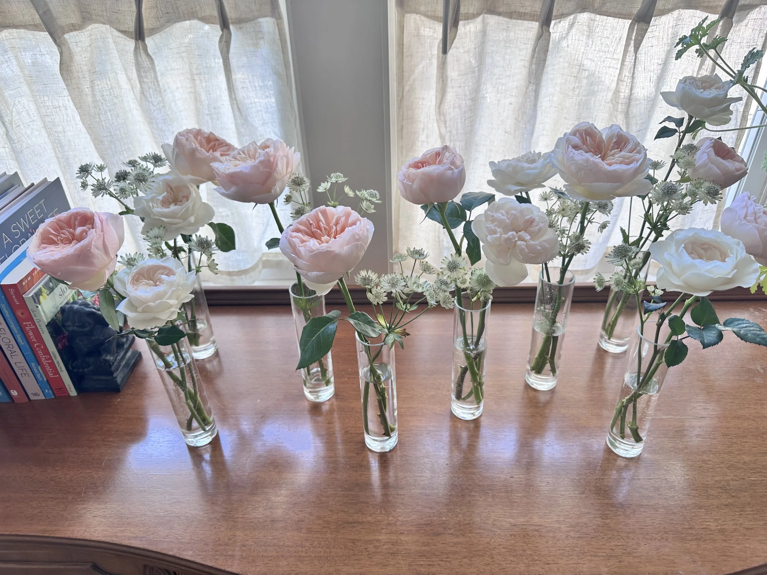 A row of glass vases filled with pink and white roses and small white flowers, placed on a wooden surface in front of a window with light-colored curtains.