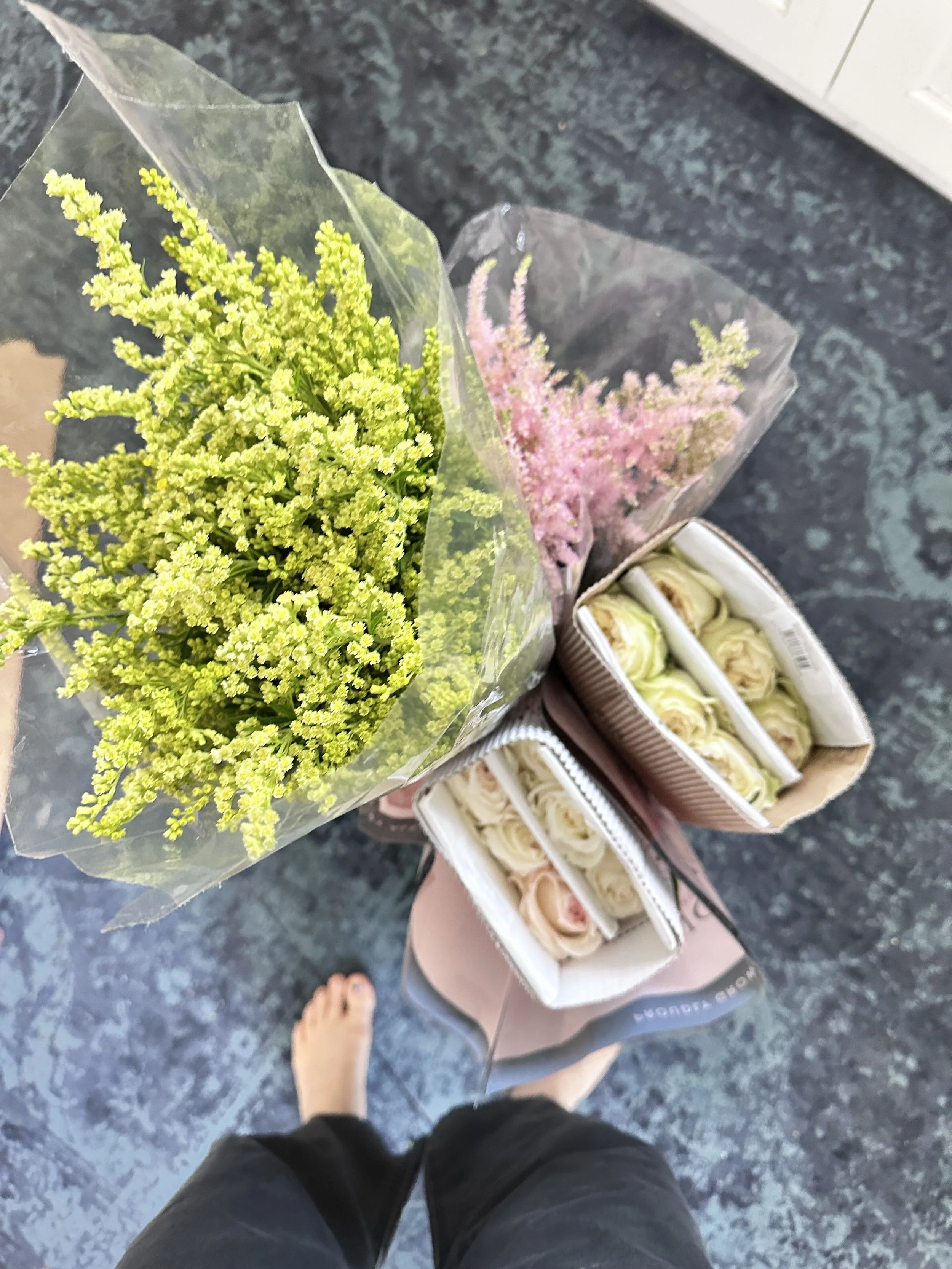 A person standing on a dark stone floor, g a bouquet of yellow and pink flowers wrapped in clear plastic, and unboxed Alexandra Farms Garden Roses in Greenwich, Connecticut.