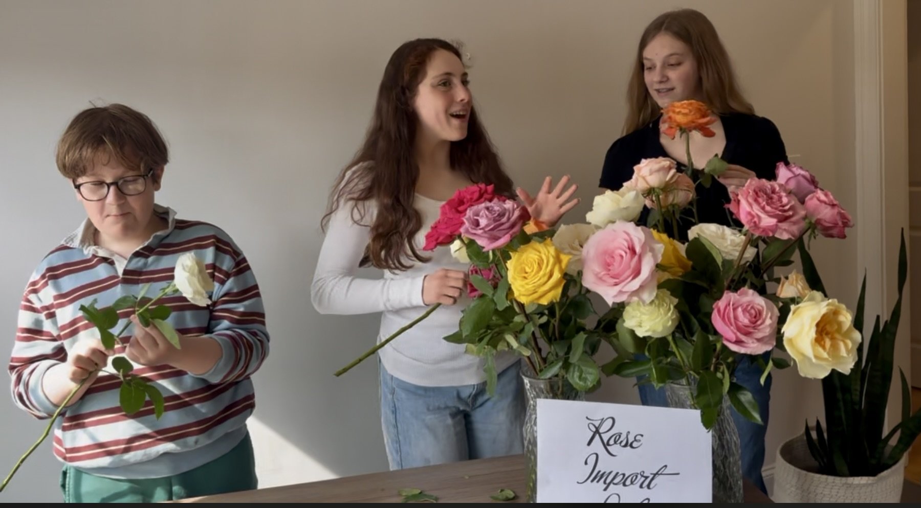 Three people at a table with a large assortment of colorful roses in a vase, with a sign that says 'Rose Import' in front of them. The person on the left, a boy with glasses, holds a white rose. The woman in the middle has curly hair and appears excited, while the girl on the right looks at the flowers with interest.