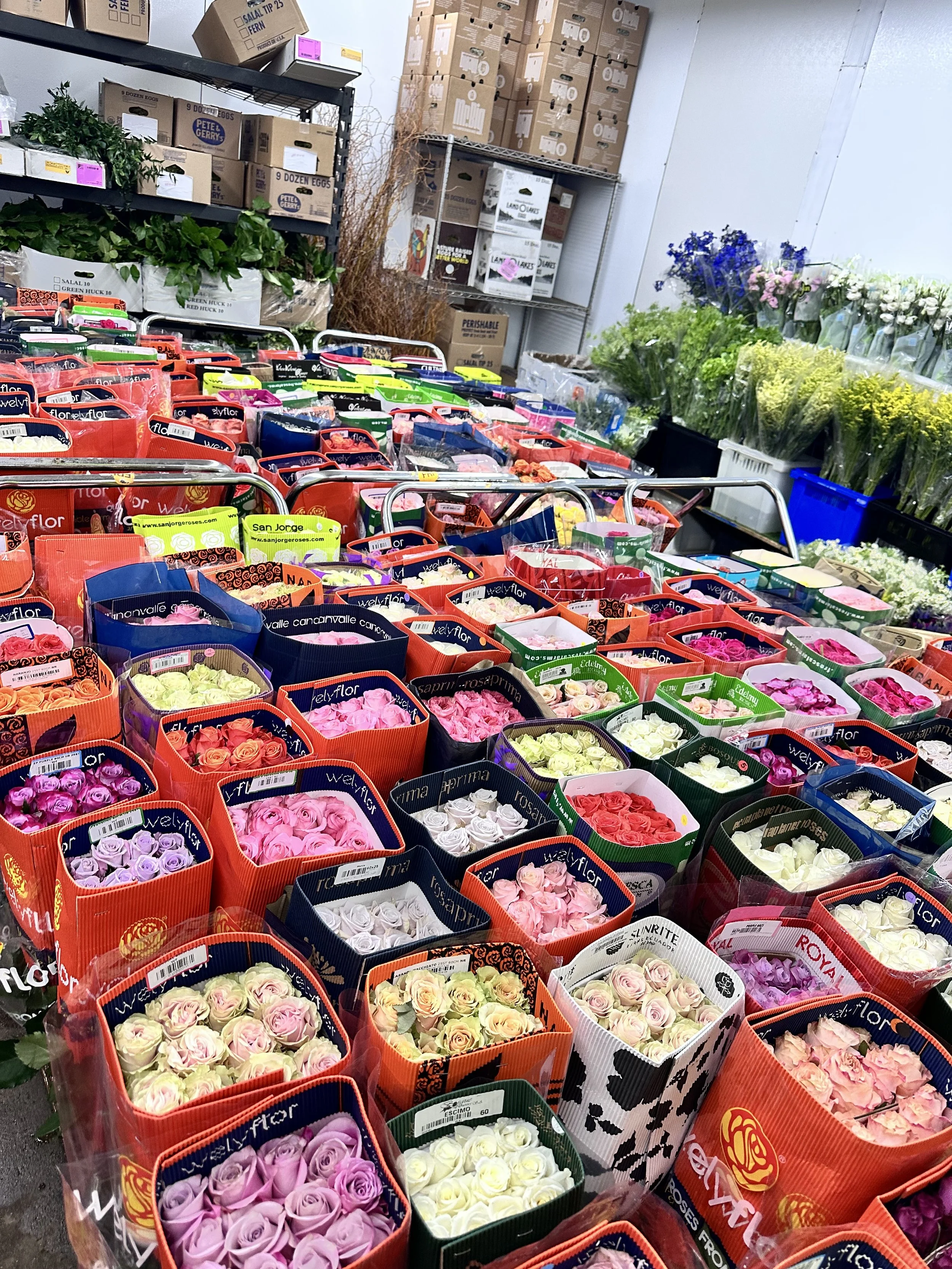 Display of various colored roses in retail shopping bags at a flower wholesale, with boxes and shelves in the background.