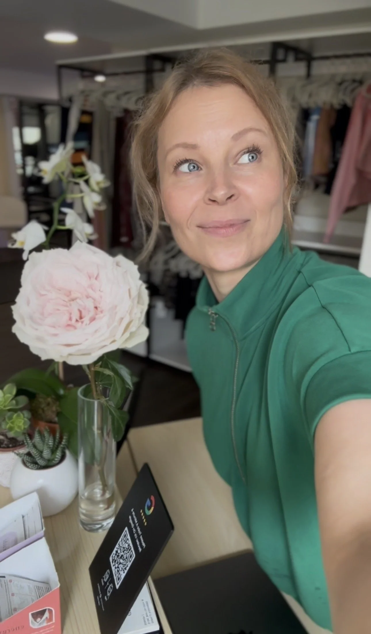 A woman with light skin, blonde hair, and blue eyes taking a selfie in an indoor setting. She is wearing a green zip-up top. In front of her are potted plants and a QR code display on a table.