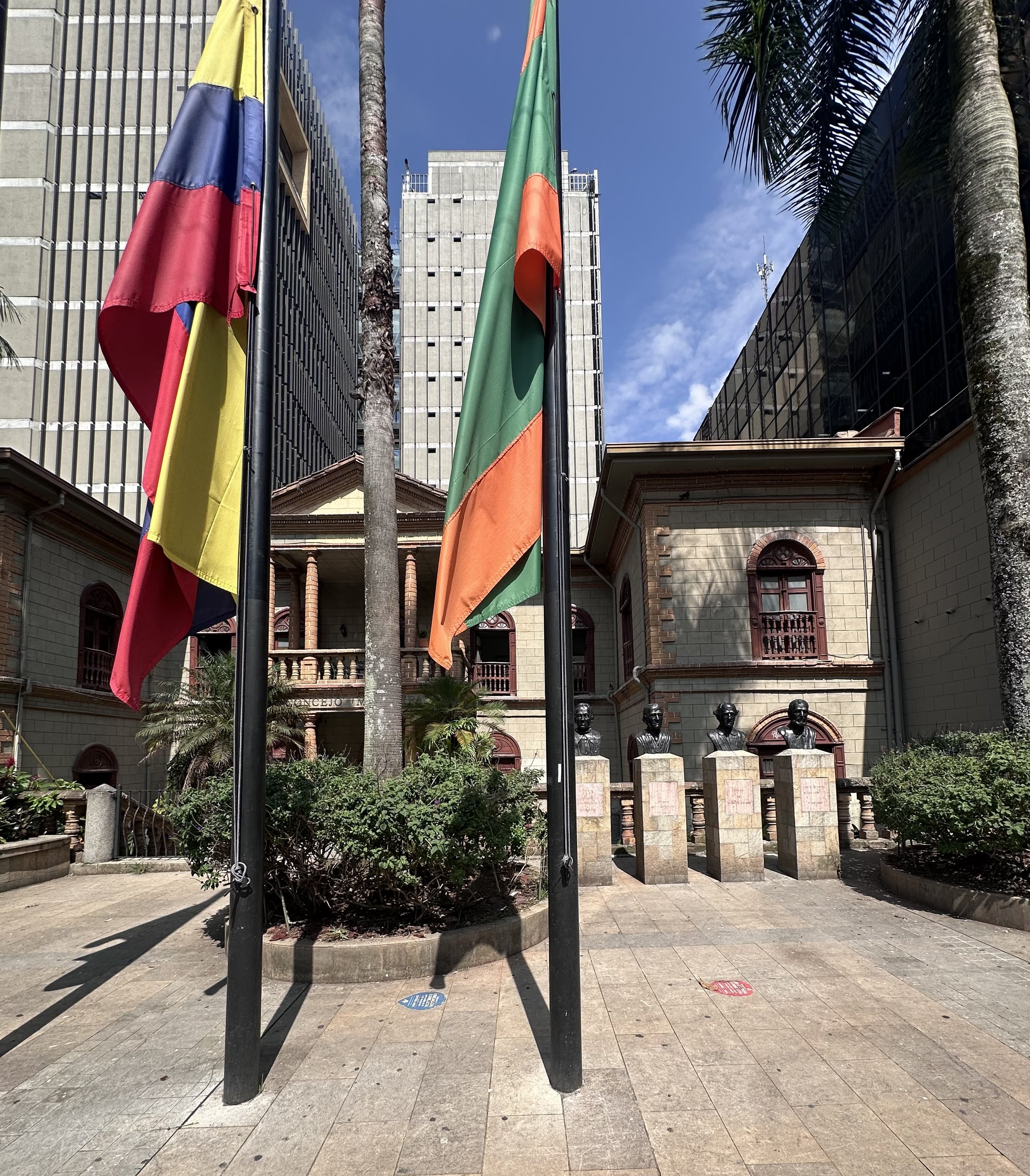 View of a city square with flagpoles, one displaying the Colombian and Envigado flags, and a historical building with statues in front. Modern skyscrapers are in the background under a partly cloudy sky. This photo was taken in Envigado, Colombia.