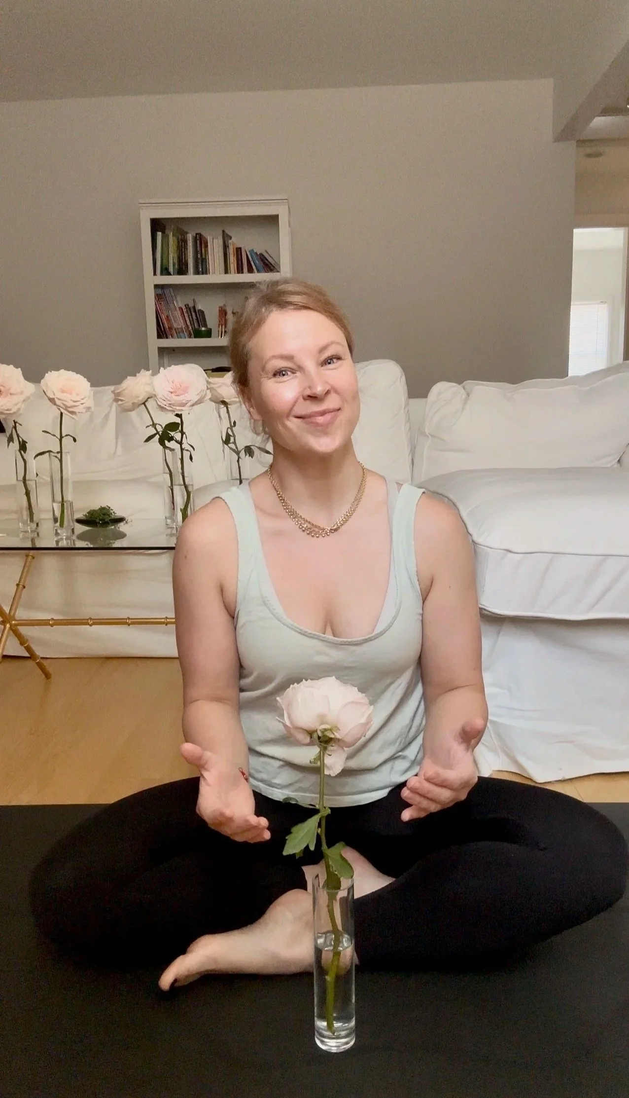 Woman sitting cross-legged on the floor holding a pink garden rose in a glass vase, with similar garden roses on a table behind her in a living room.