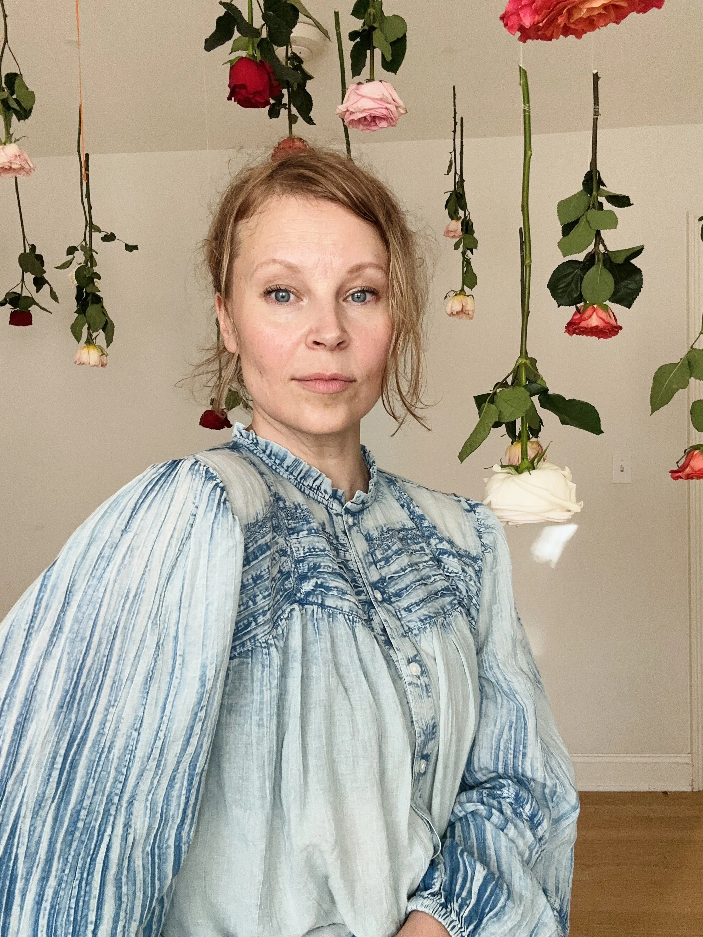 A woman with light brown curly hair wearing a blue and white striped shirt. She is indoors with hanging garden roses (red, pink, and white) above her head and a plain white wall behind her.
