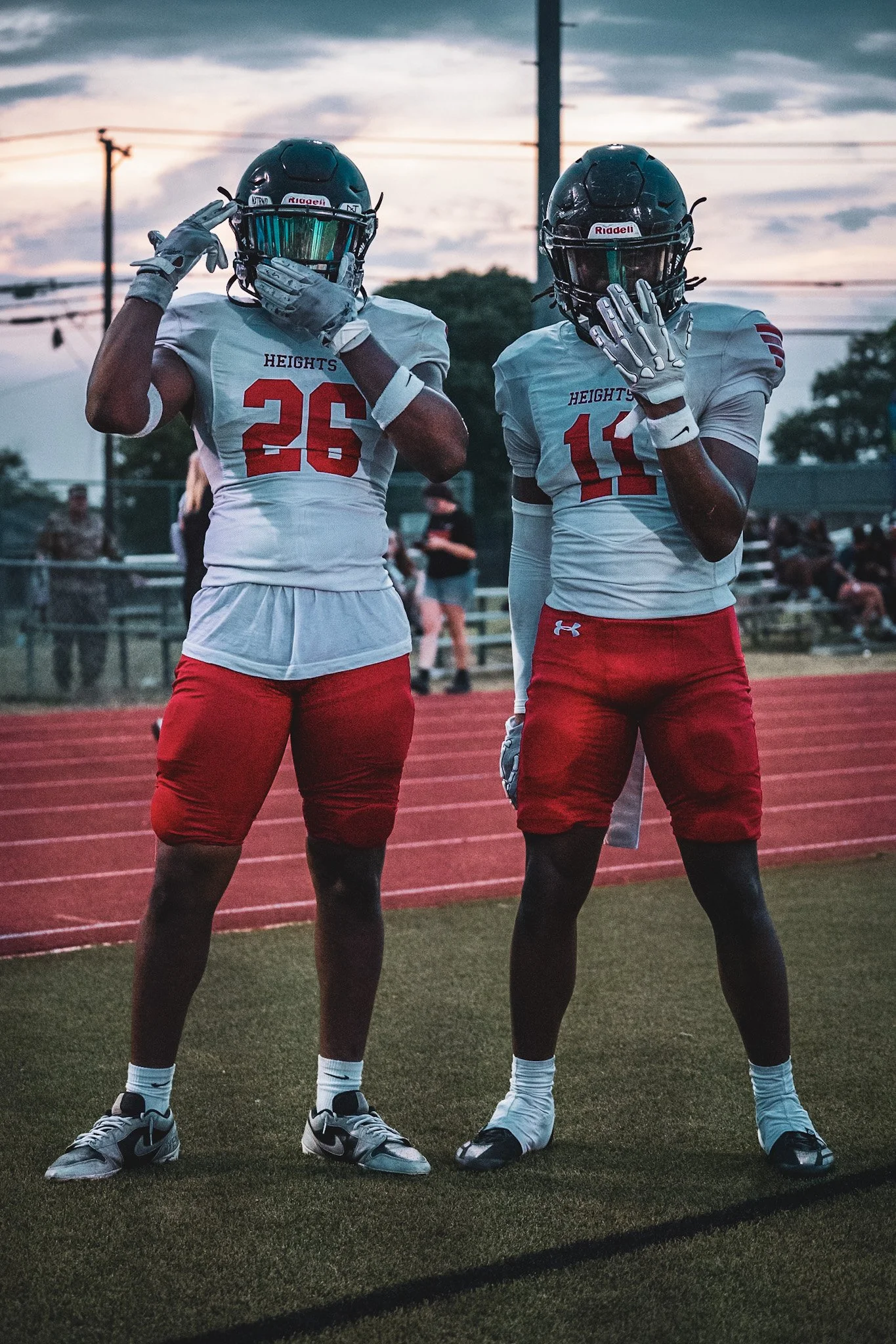 Two football players in white jerseys and red shorts standing on a track, wearing helmets and gloves, with their hands near their faces, during sunset at a stadium.