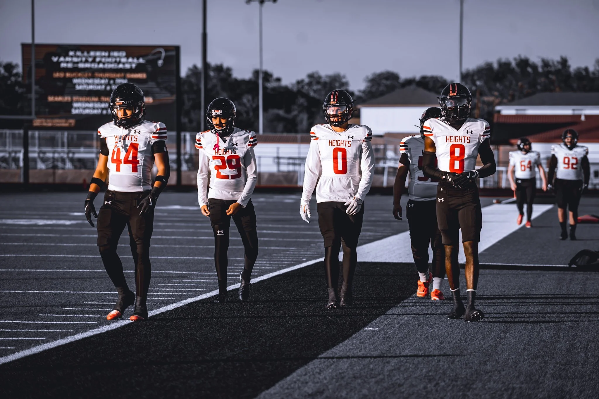 Young football players in white jerseys with red numbers, black pants, and helmets walk along a football field during dusk.