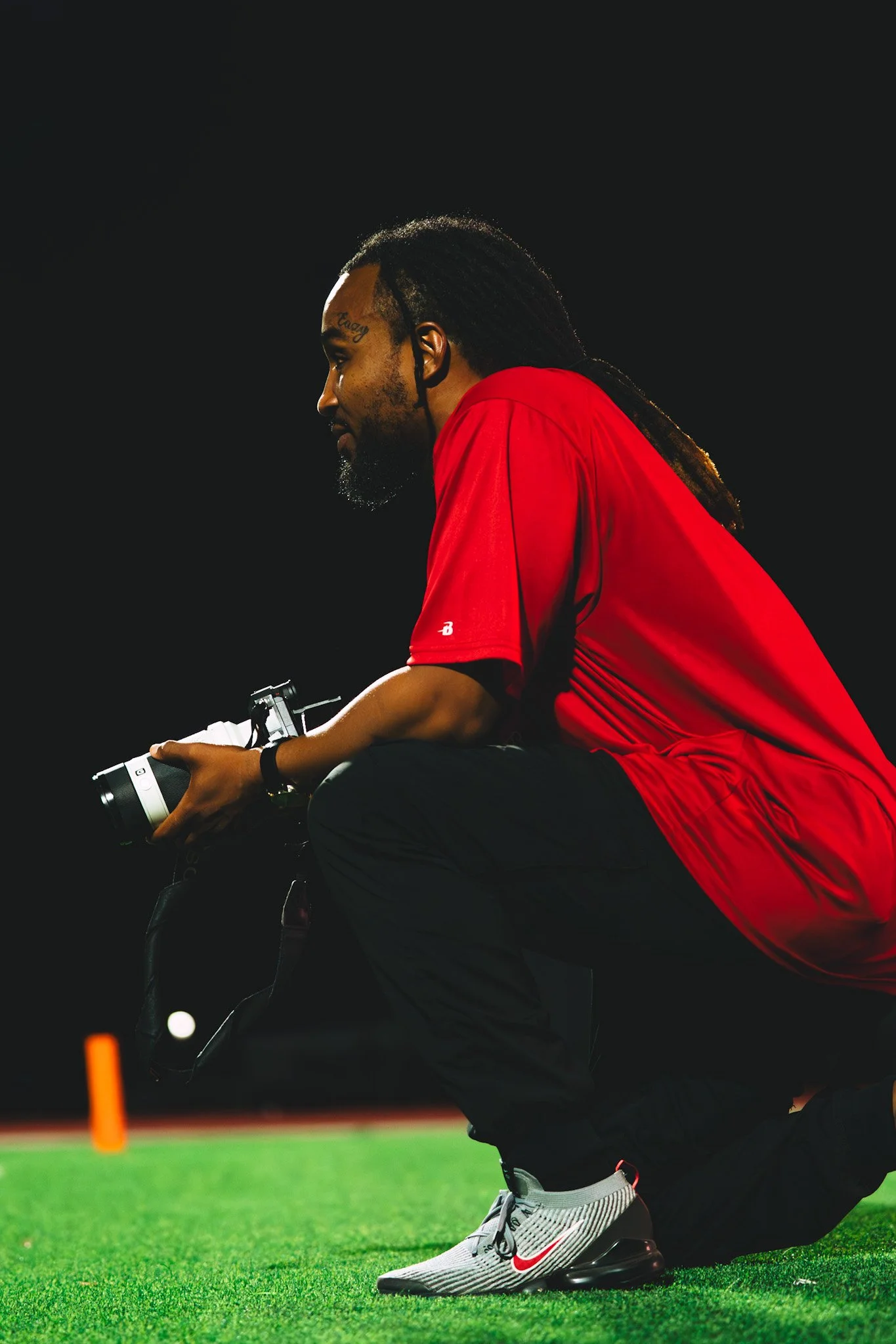 A man wearing a red shirt, black pants, and Nike sneakers crouches on a football field at night, holding a camera with a black strap.