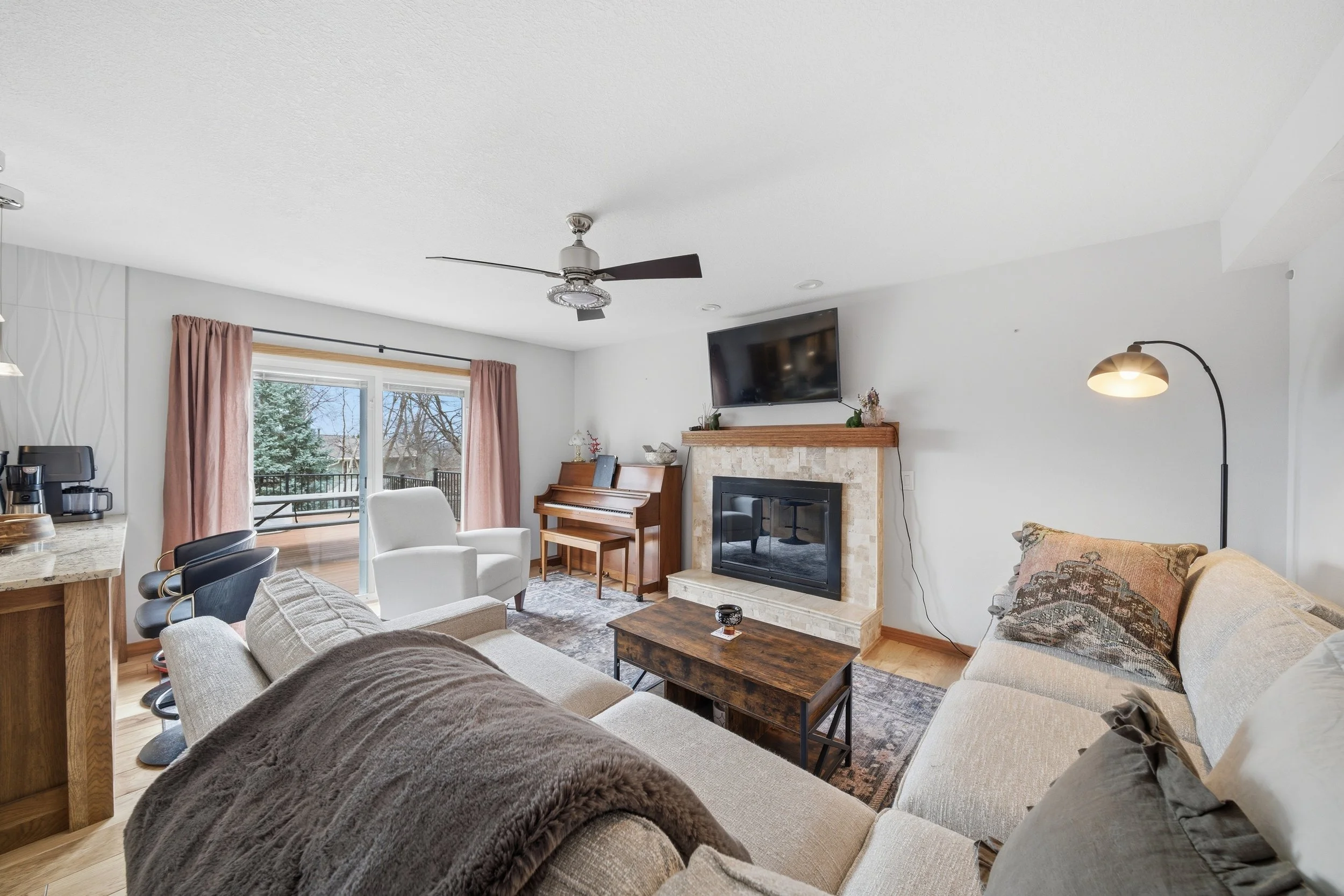 Real estate photography of a living room with beige sofas, a fireplace with a mounted TV above, a wooden coffee table, a white armchair near sliding glass doors with pink curtains, and a ceiling fan.