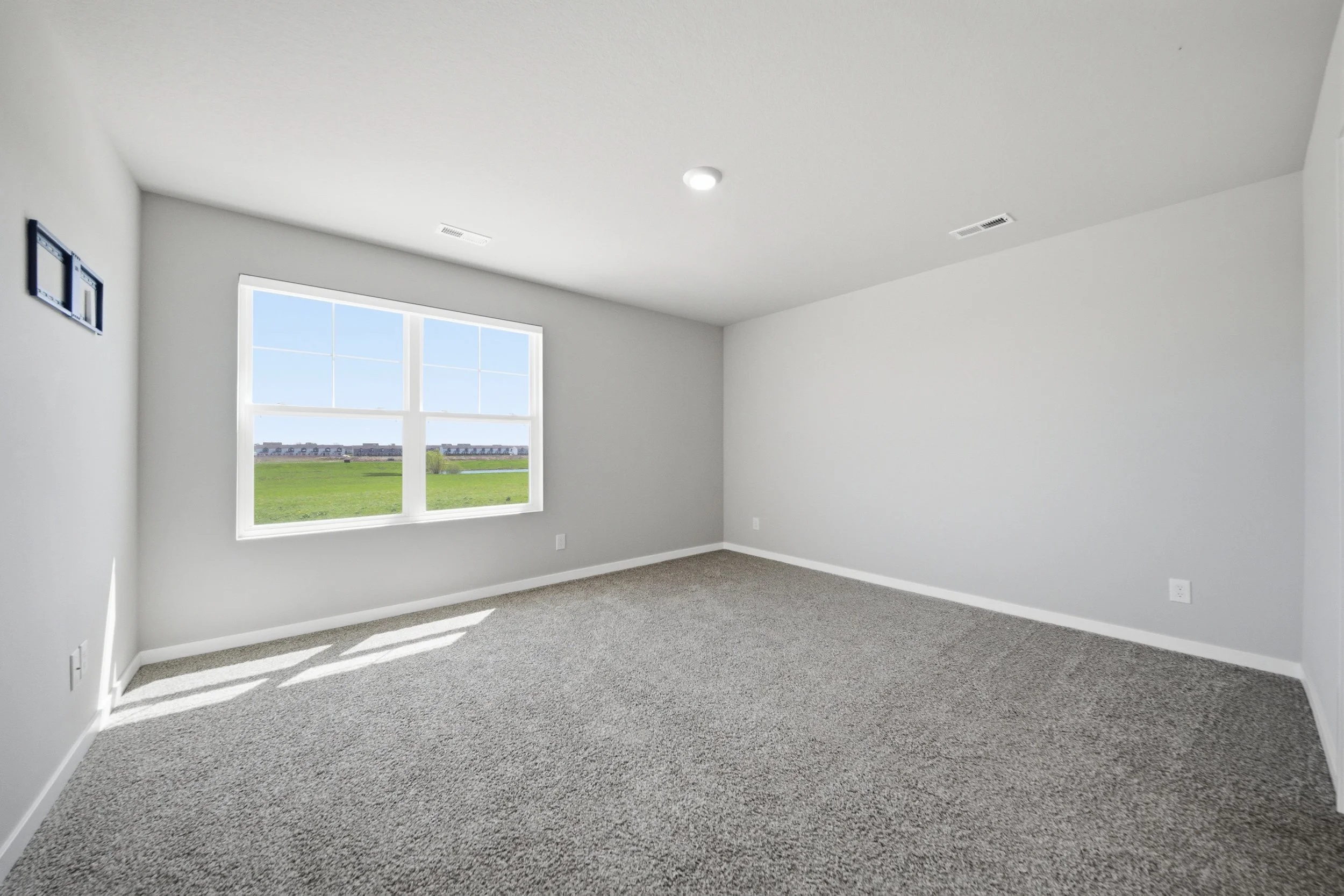 Real estate photography of an empty room with large window showing green field and blue sky, gray carpet, white walls, ceiling, and vent.