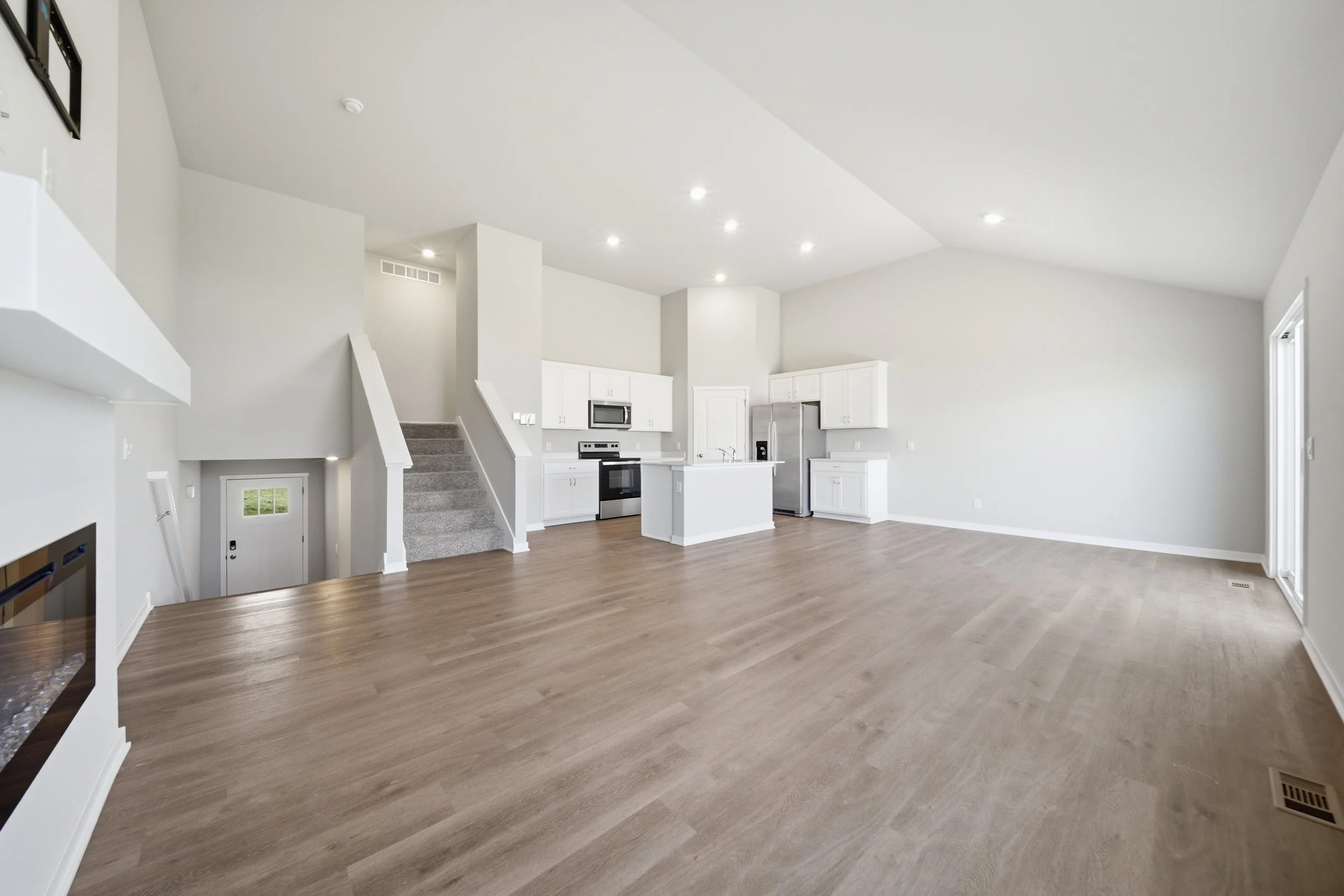 Real estate photography of an empty living room and kitchen area in a modern house with white walls, wood flooring, and recessed lighting.