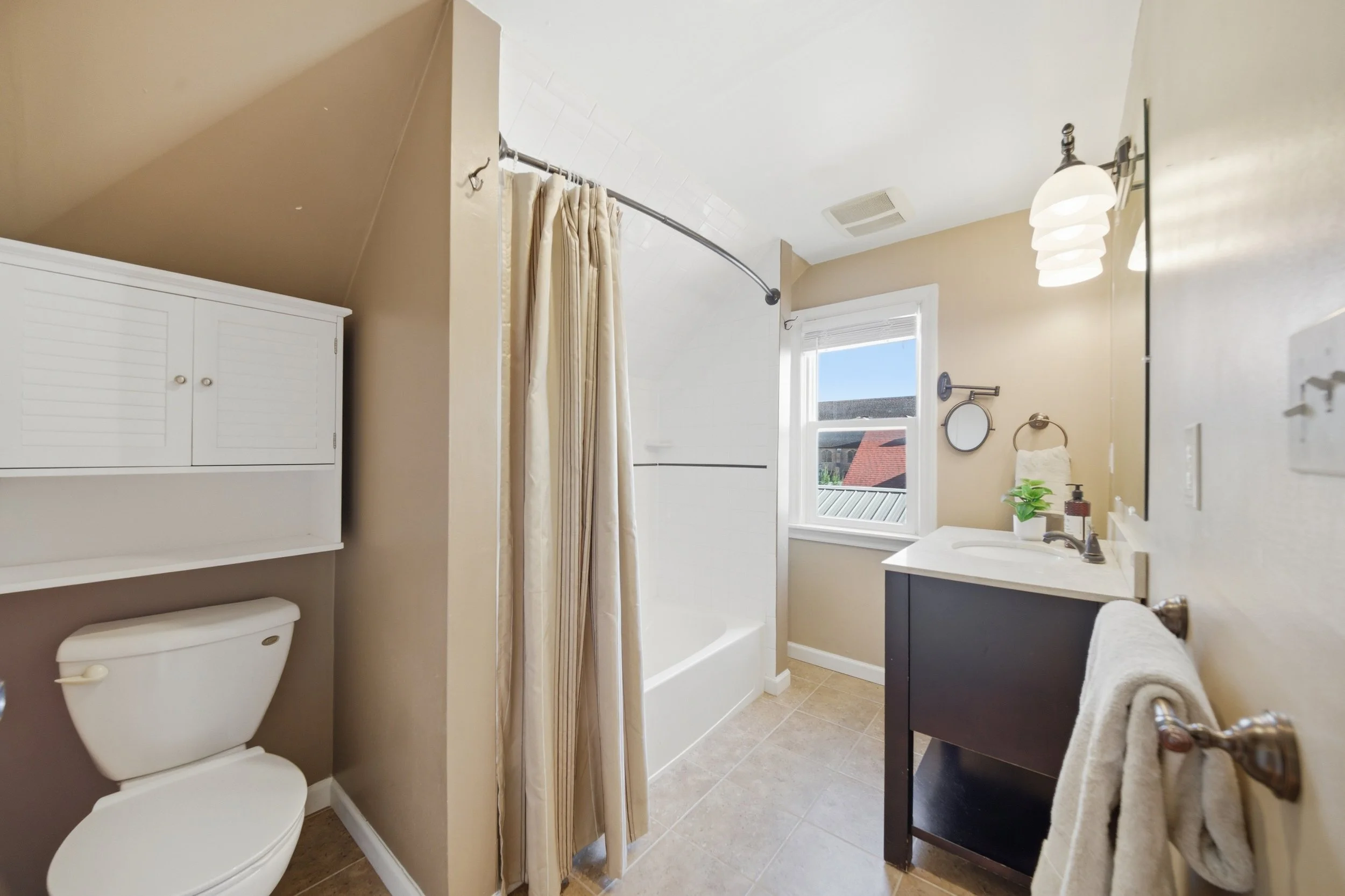 Real estate photography of a bathroom with a toilet, a shower with a beige curtain, a window letting in natural light, a dark vanity with a white sink, a towel rack with a towel, and a decorative mirror with three lights above it.