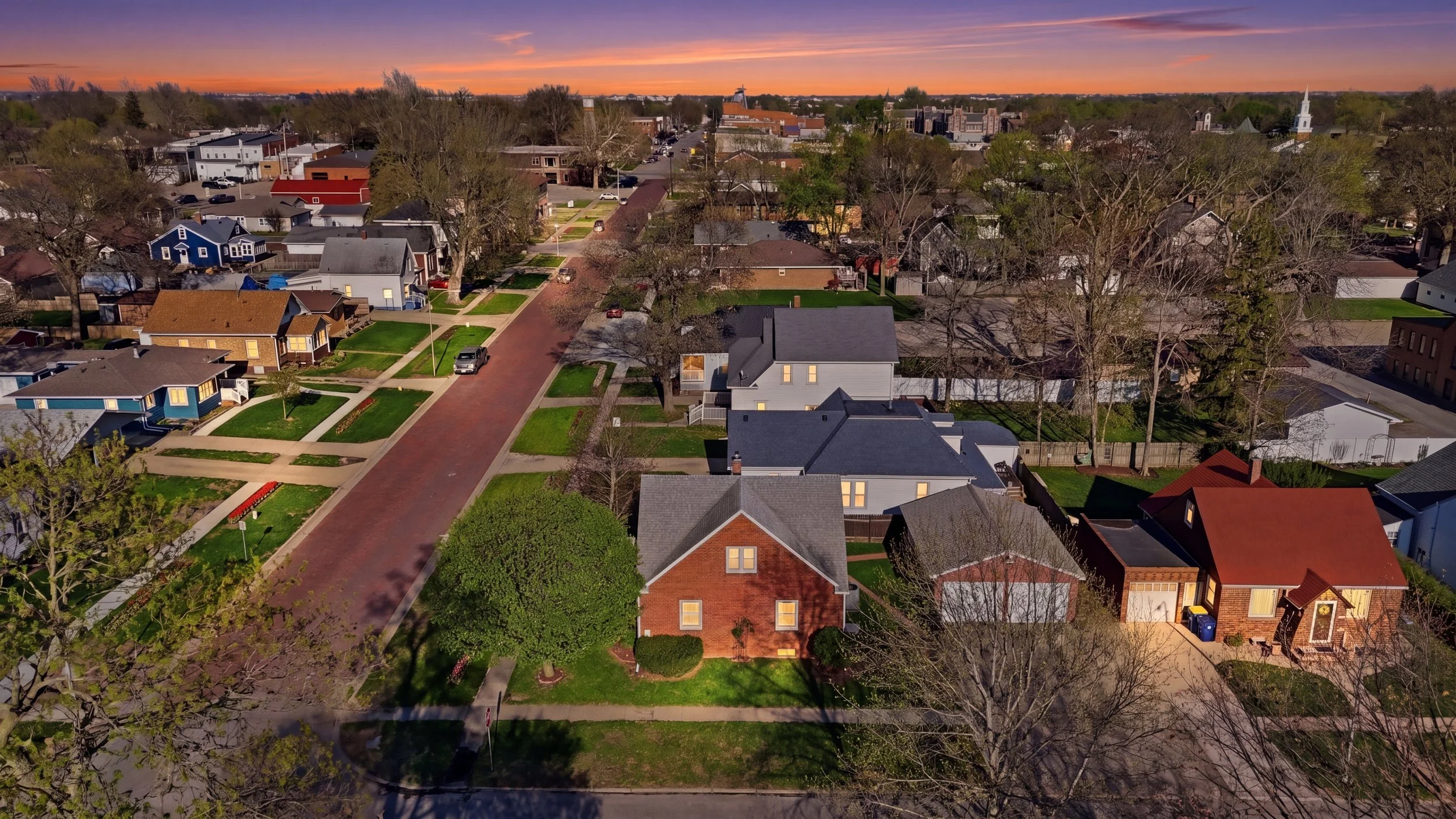 Real estate virtual twilight photography of an aerial view of a residential neighborhood at sunset, showing a variety of houses, trees, and a street running through the community.