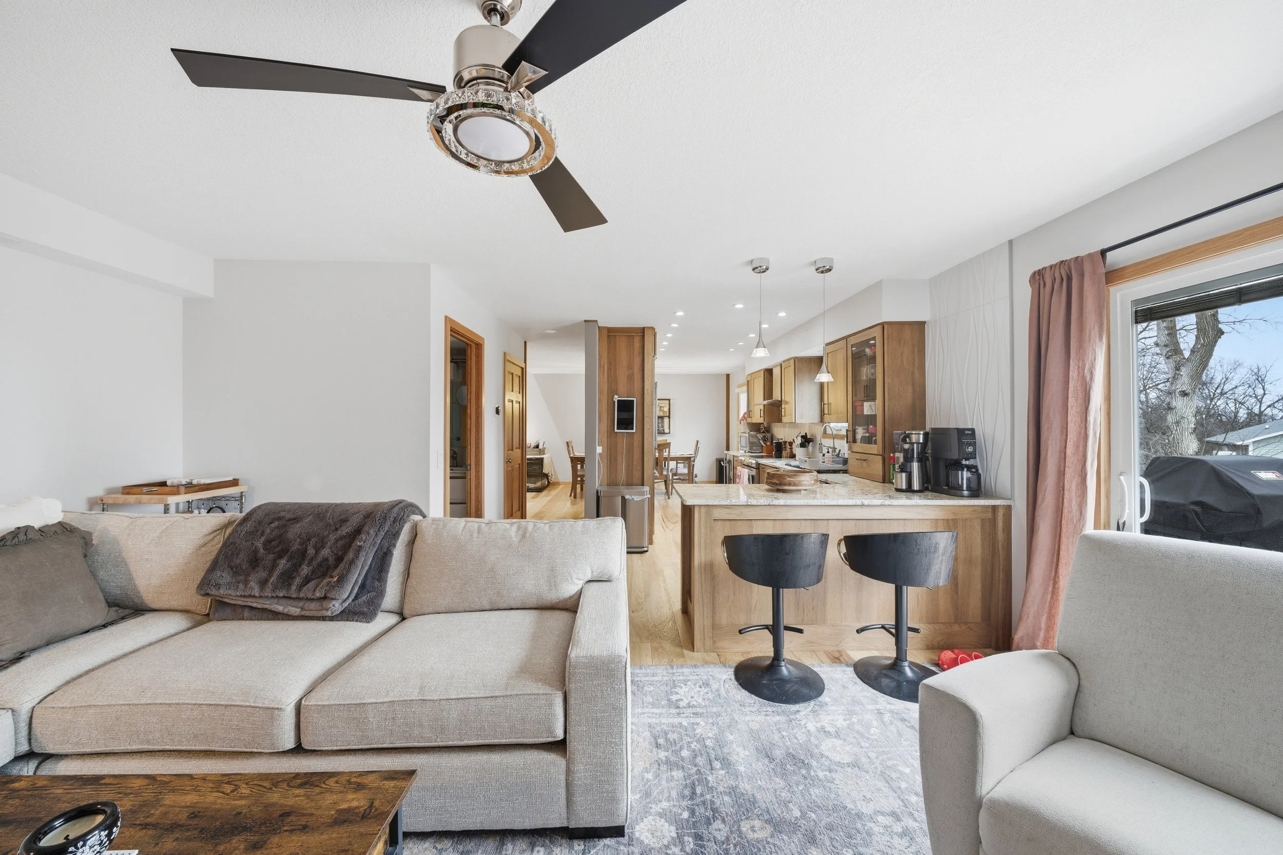 Real estate photography of a living room and kitchen area with beige sofa, black chairs at the island, white walls, wooden cabinets, kitchen appliances, and sliding glass door with pink curtains.