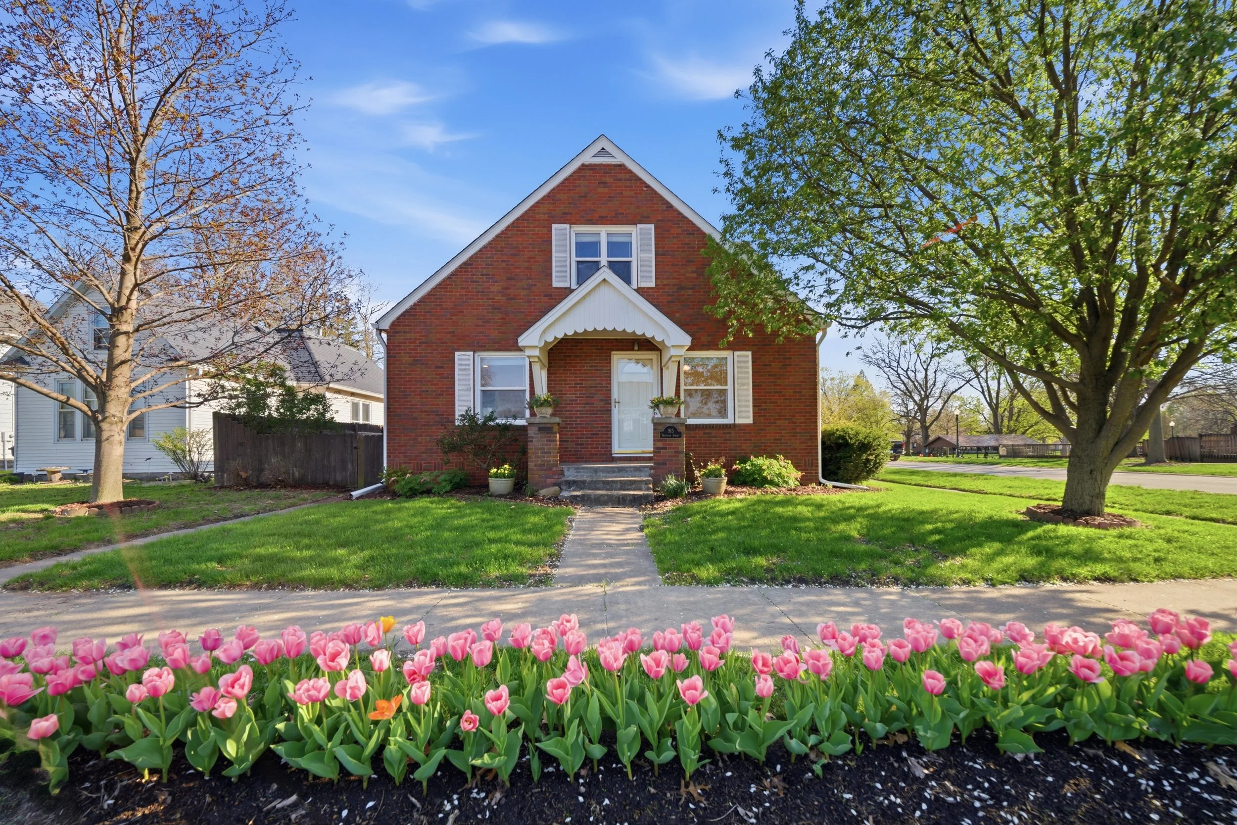 Real estate photography of a brick house with white shutters and a white front door, surrounded by a green lawn, trees, and a flowerbed with pink tulips in the foreground.