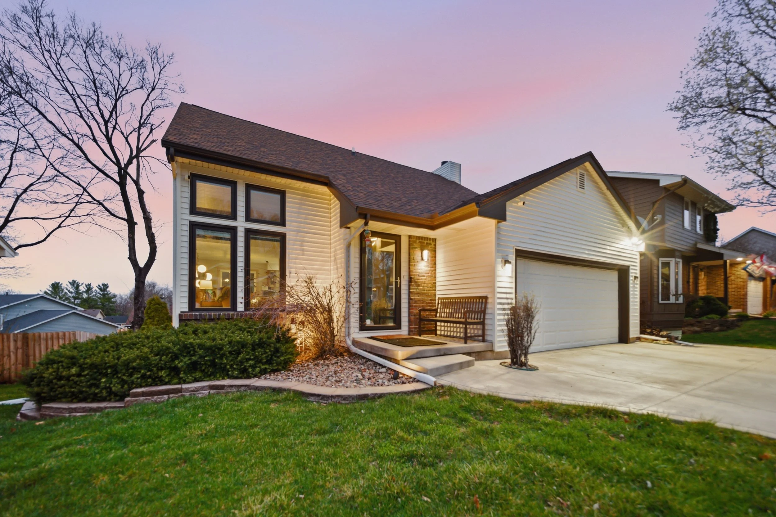 Front view of a modern two-story house with white siding, brick accents, and a brown shingle roof during sunset, with a green lawn, some bushes, and a large tree in the background.