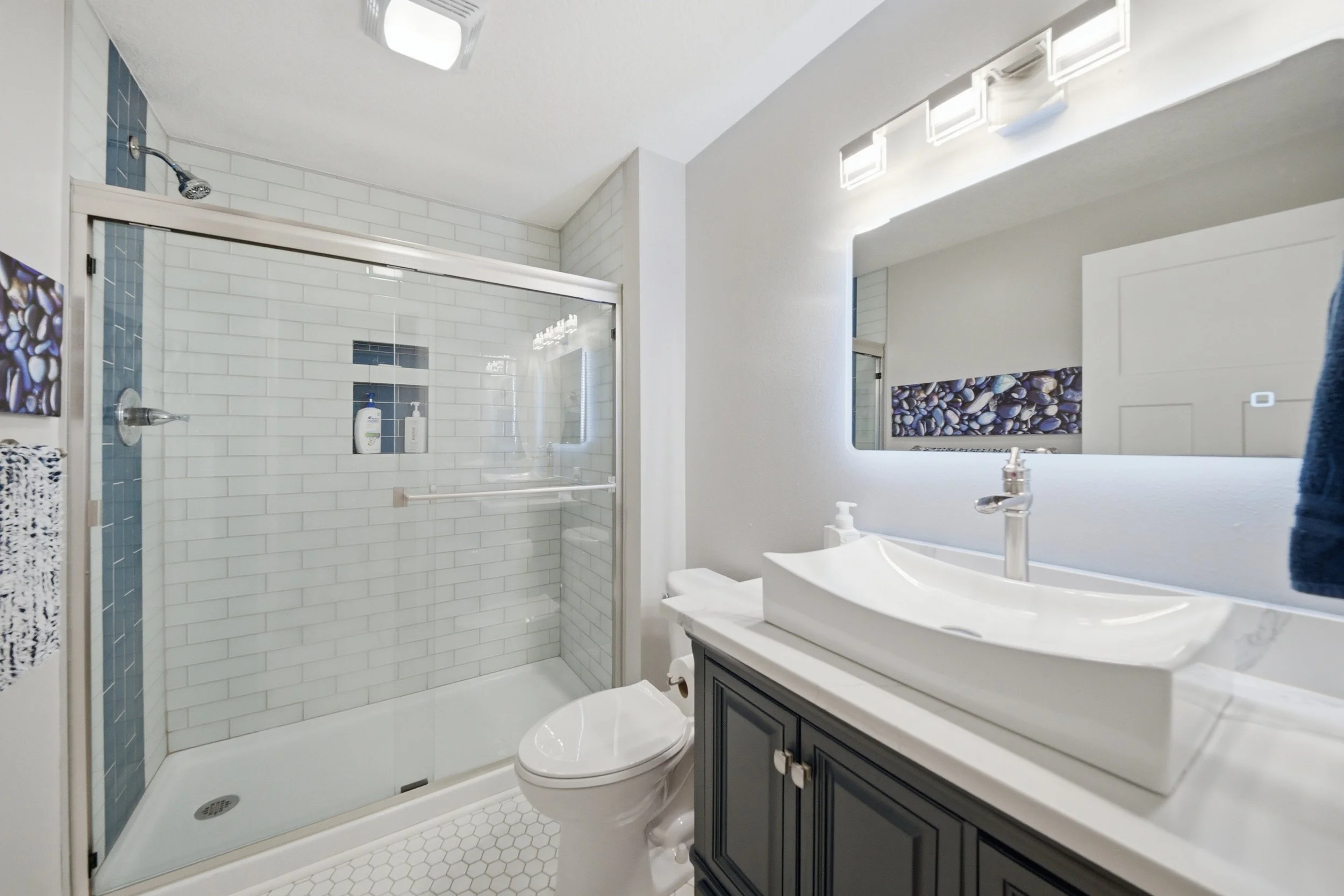 Bathroom with a glass shower enclosure, white subway tile walls with a vertical strip of small blue tiles, a white sink with a gray vanity, a mirror with integrated lights, and a toilet.