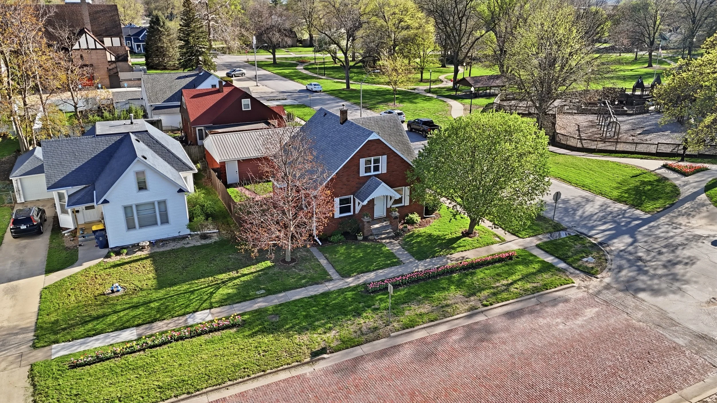 Real estate photography of an aerial view of a neighborhood with houses, a park, trees, sidewalks, and streets in spring.