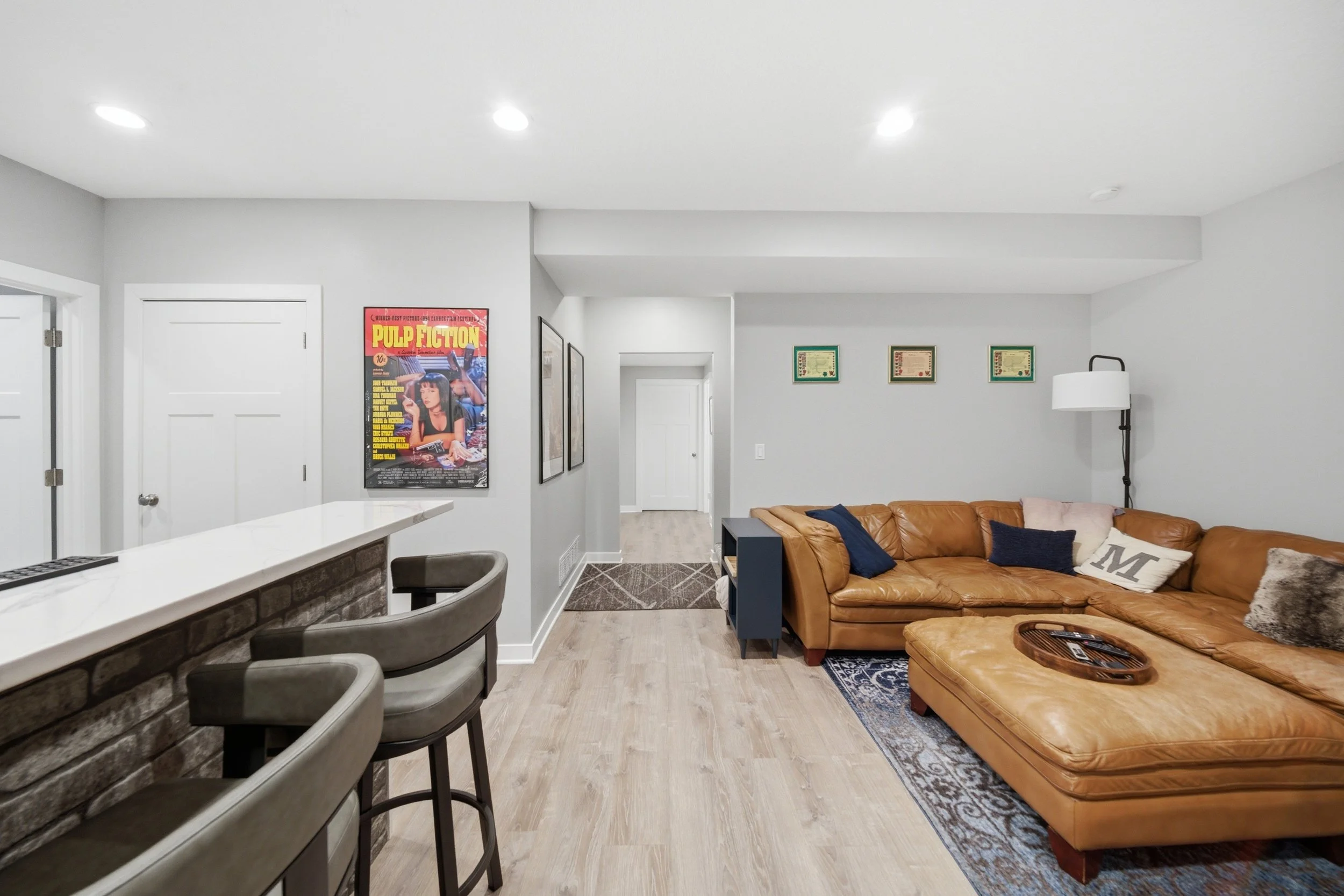 Living room with a tan leather sectional sofa, pillows, a patterned area rug, wall-mounted lamps, framed artwork, a white ceiling, and a white door leading to a hallway.