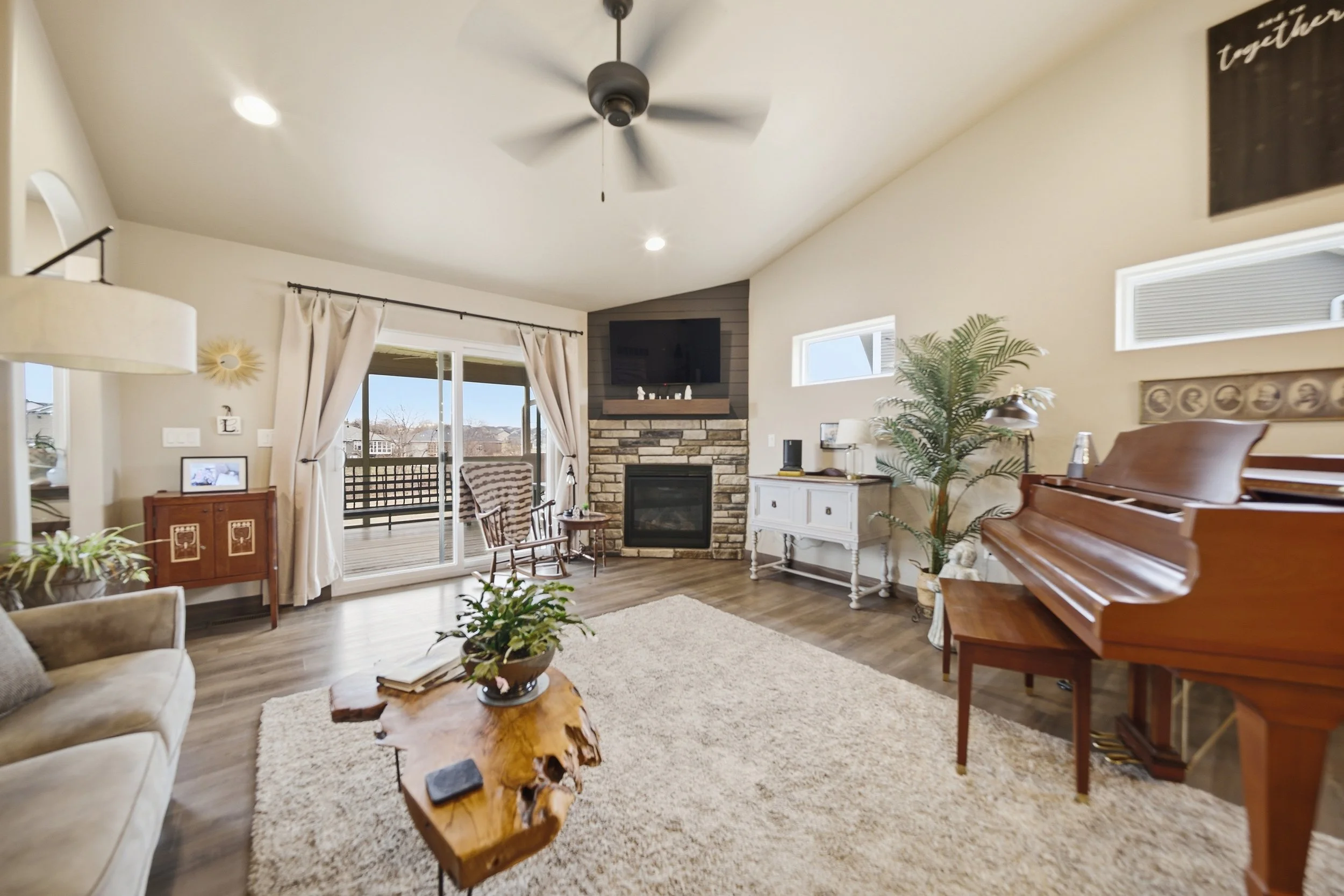 Living room with a piano on the right, a fireplace with a mounted TV above, a sliding glass door with curtains leading to a deck, various side tables, plants, a cream-colored armchair, a beige sofa, and a wooden coffee table.