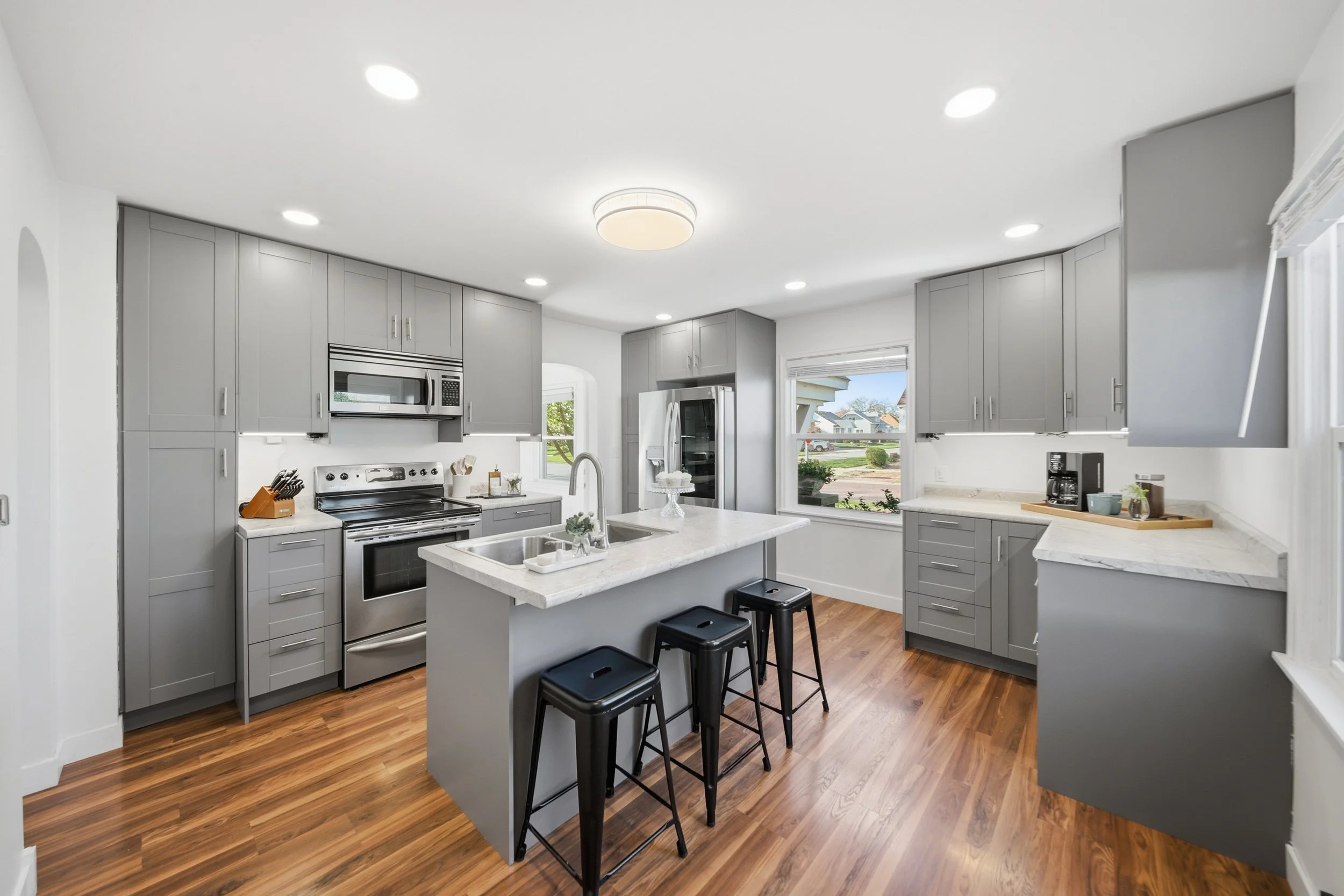 Real estate photography of a modern kitchen with gray cabinets, stainless steel appliances, a white island with stools, and hardwood floors.