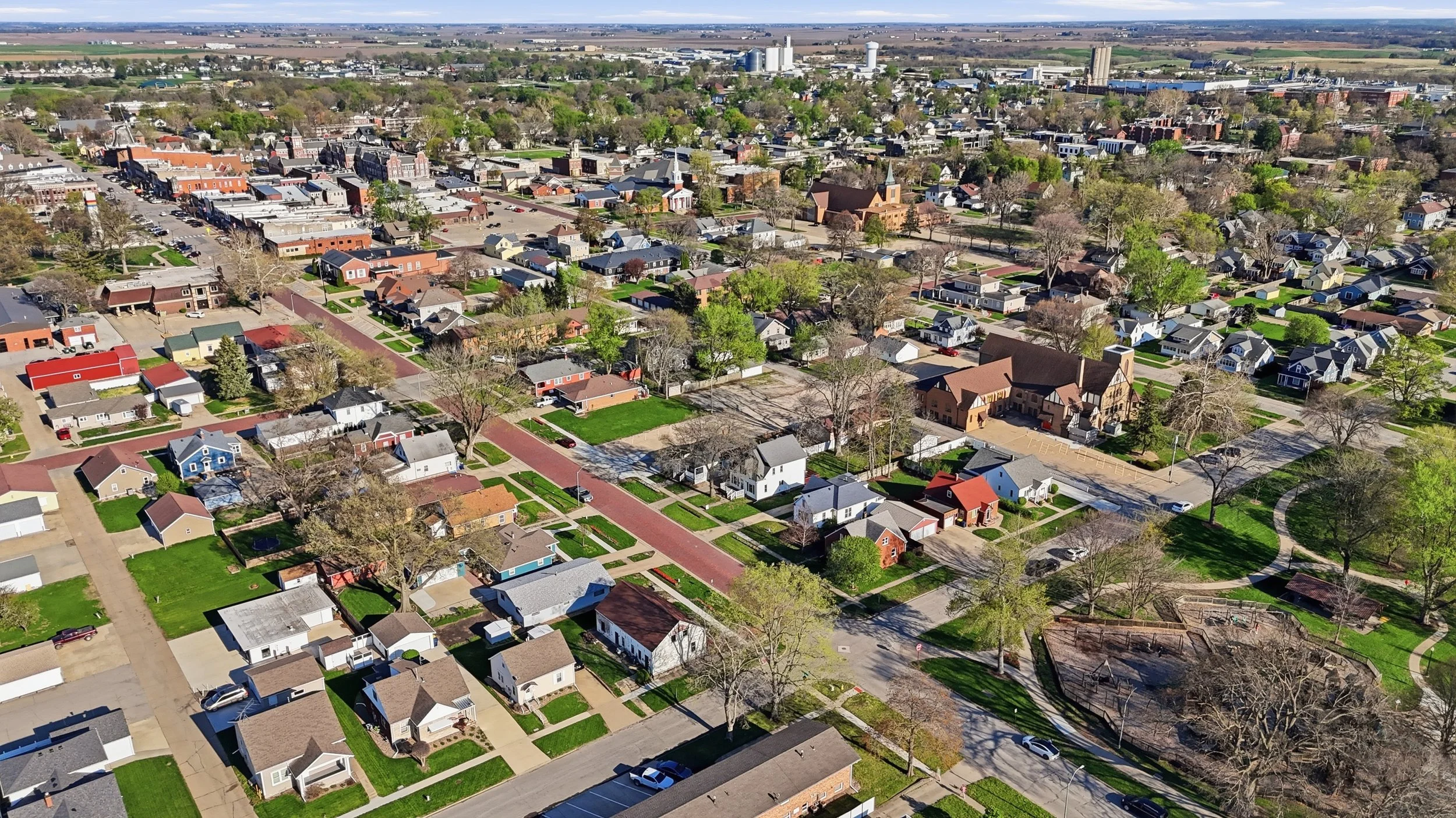 Aerial view of a suburban neighborhood with houses, trees, streets, and a downtown area in the background under a partly cloudy sky.