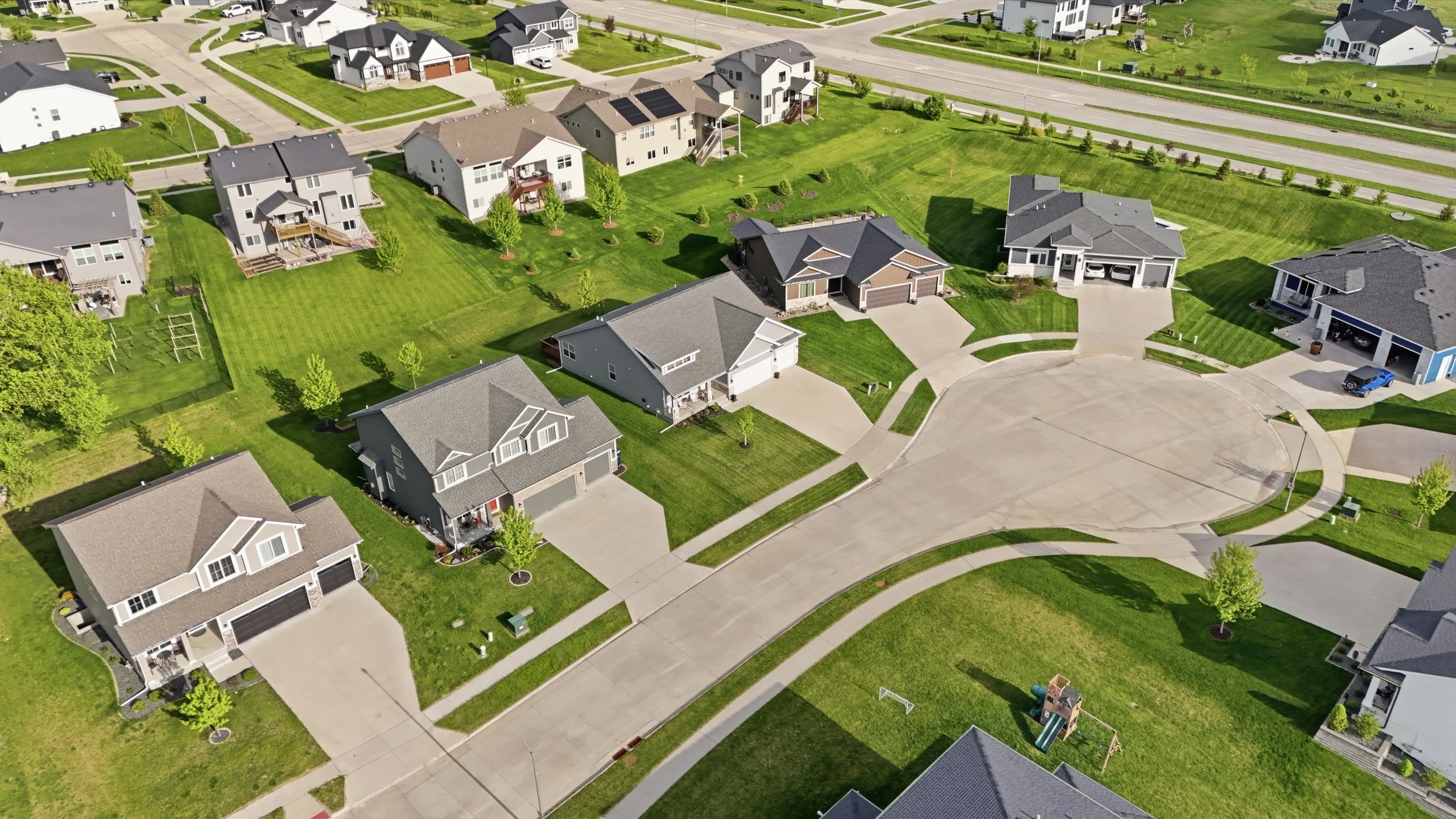 Aerial view of a suburban neighborhood with single-family homes, green lawns, trees, and wide streets.