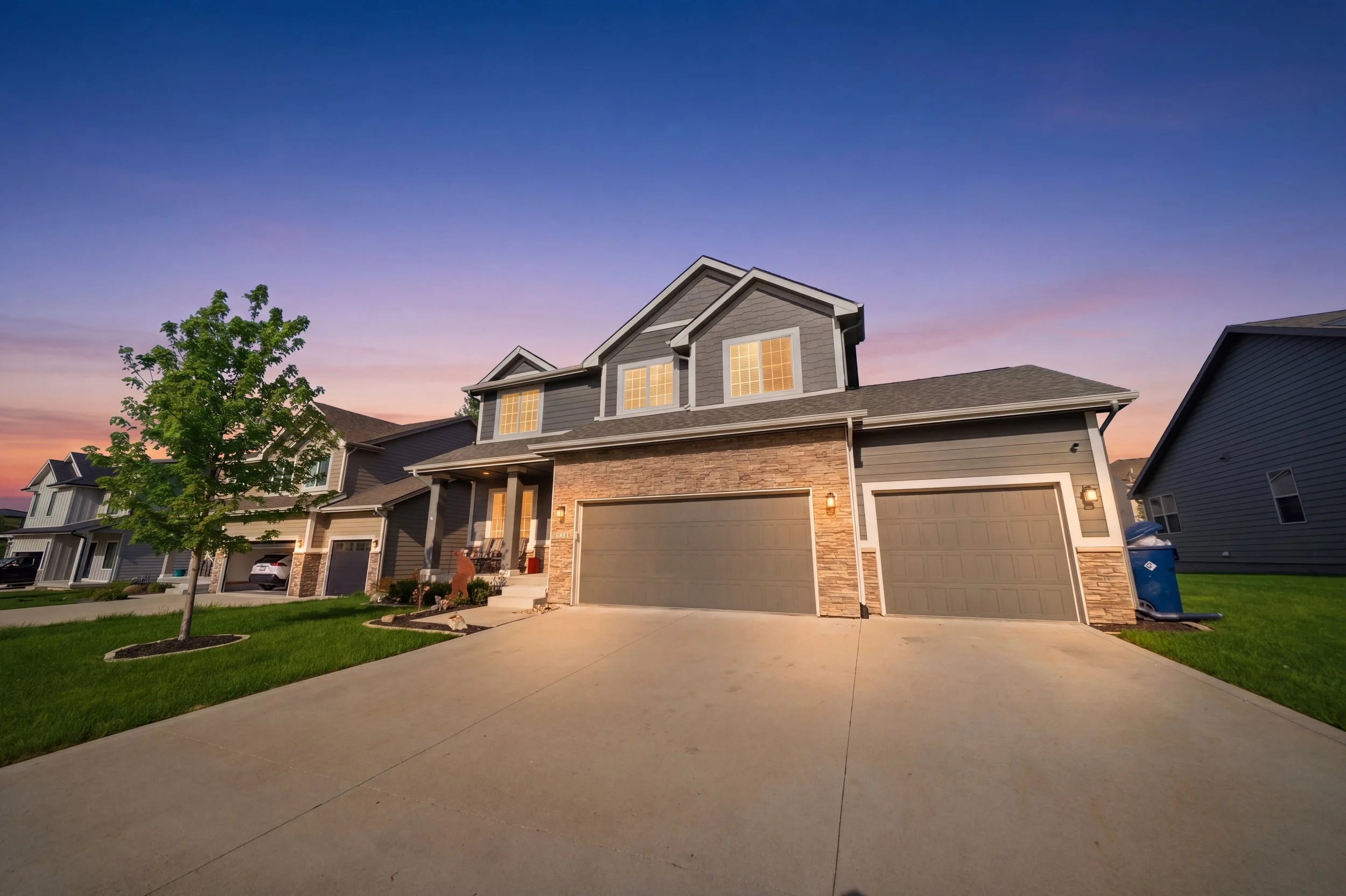 Twilight real estate photography of a a neighborhood with modern houses, a driveway, a green lawn, and a sunset sky in the background.