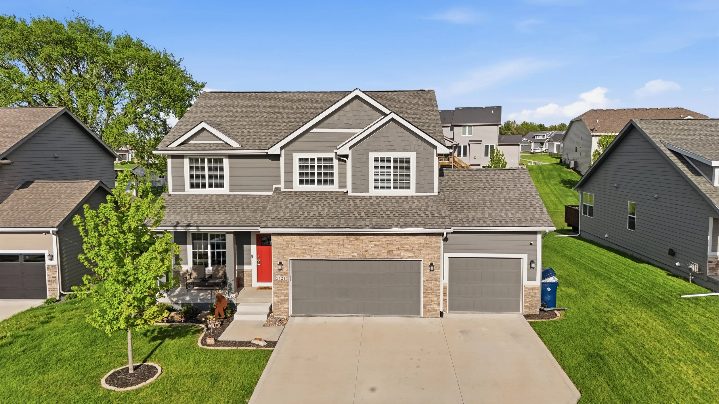 Front view of a modern, two-story suburban house with gray siding, a brick facade, and a three-car garage. The house has a landscaped front yard with a concrete driveway, a small tree, and a garden bed. Several neighboring houses are visible in the b
