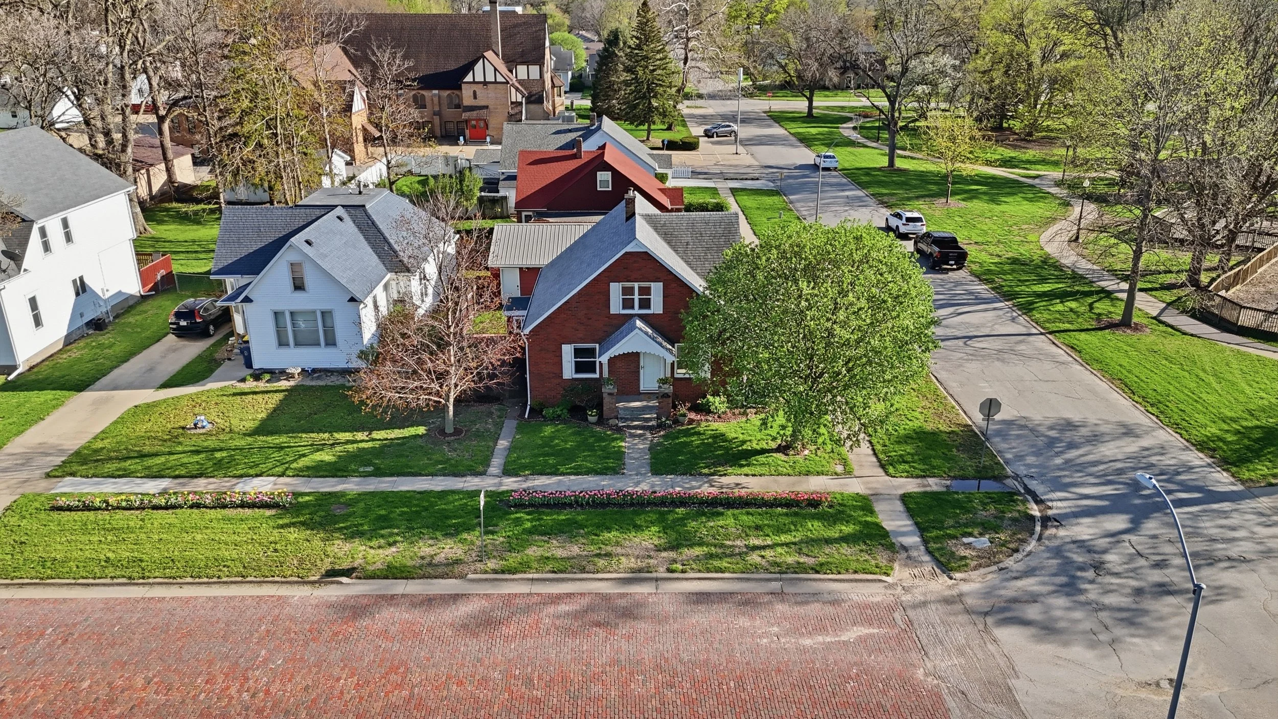 Aerial view of a suburban neighborhood with houses, trees, parked cars, sidewalks, and a street.