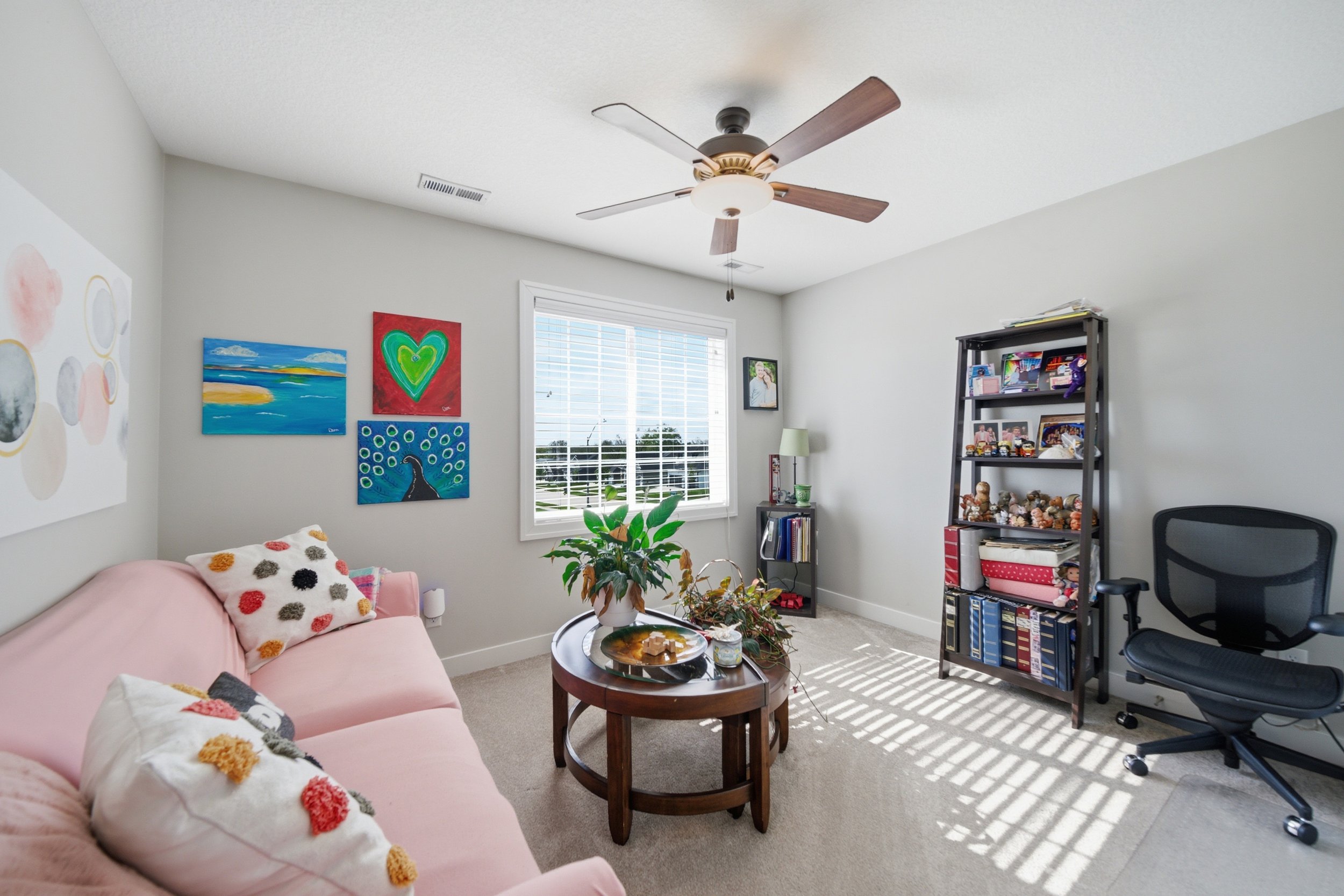 A cozy living room with a pink sofa adorned with colorful plush pillows, a wooden coffee table with decorative items, a window with white blinds letting in sunlight, colorful artwork on the walls, a black bookshelf filled with books and collectibles,