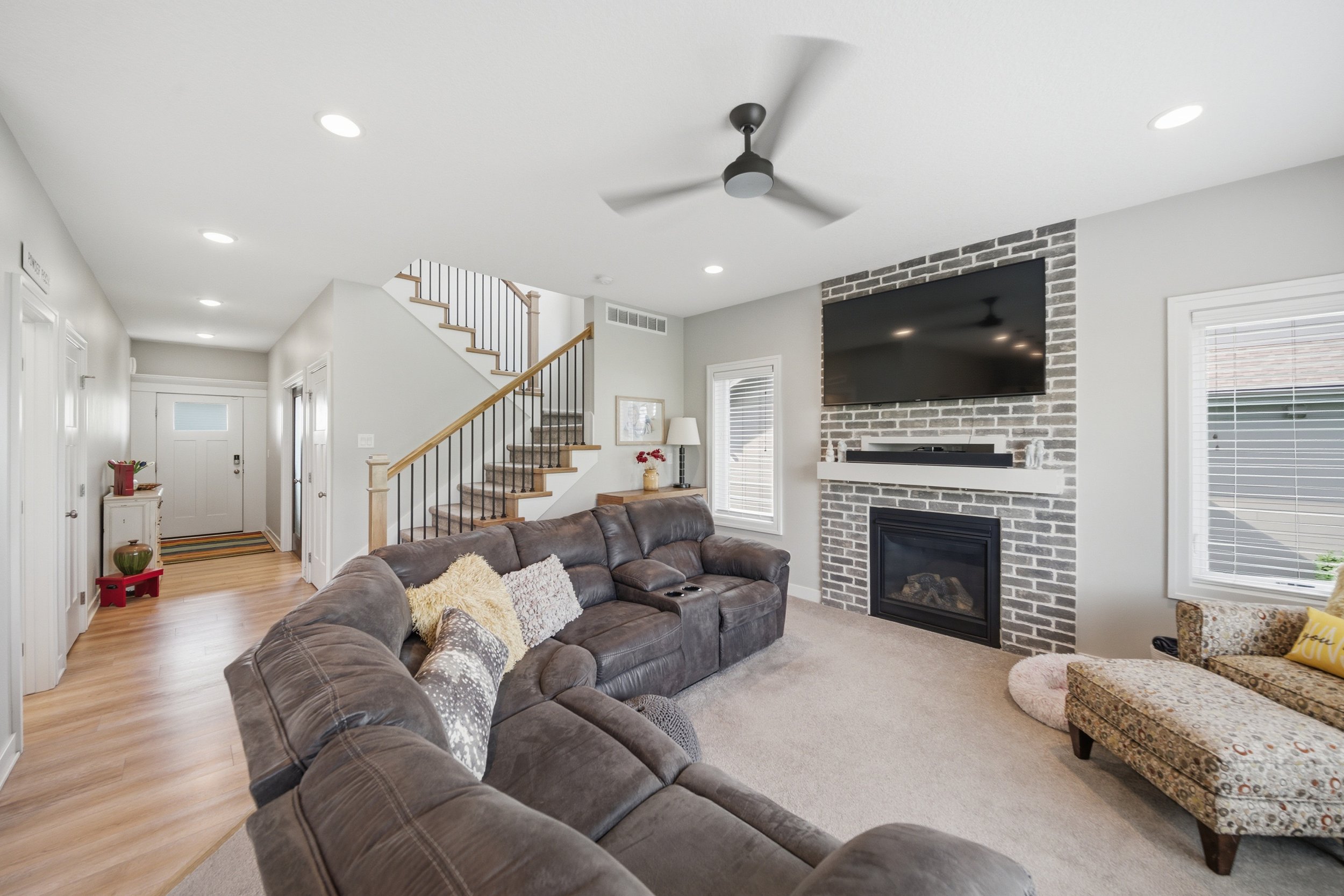 Living room with a brown leather sectional sofa, a patterned armchair, a fireplace with a mounted TV, a ceiling fan, multiple windows with blinds, and a staircase leading upstairs.