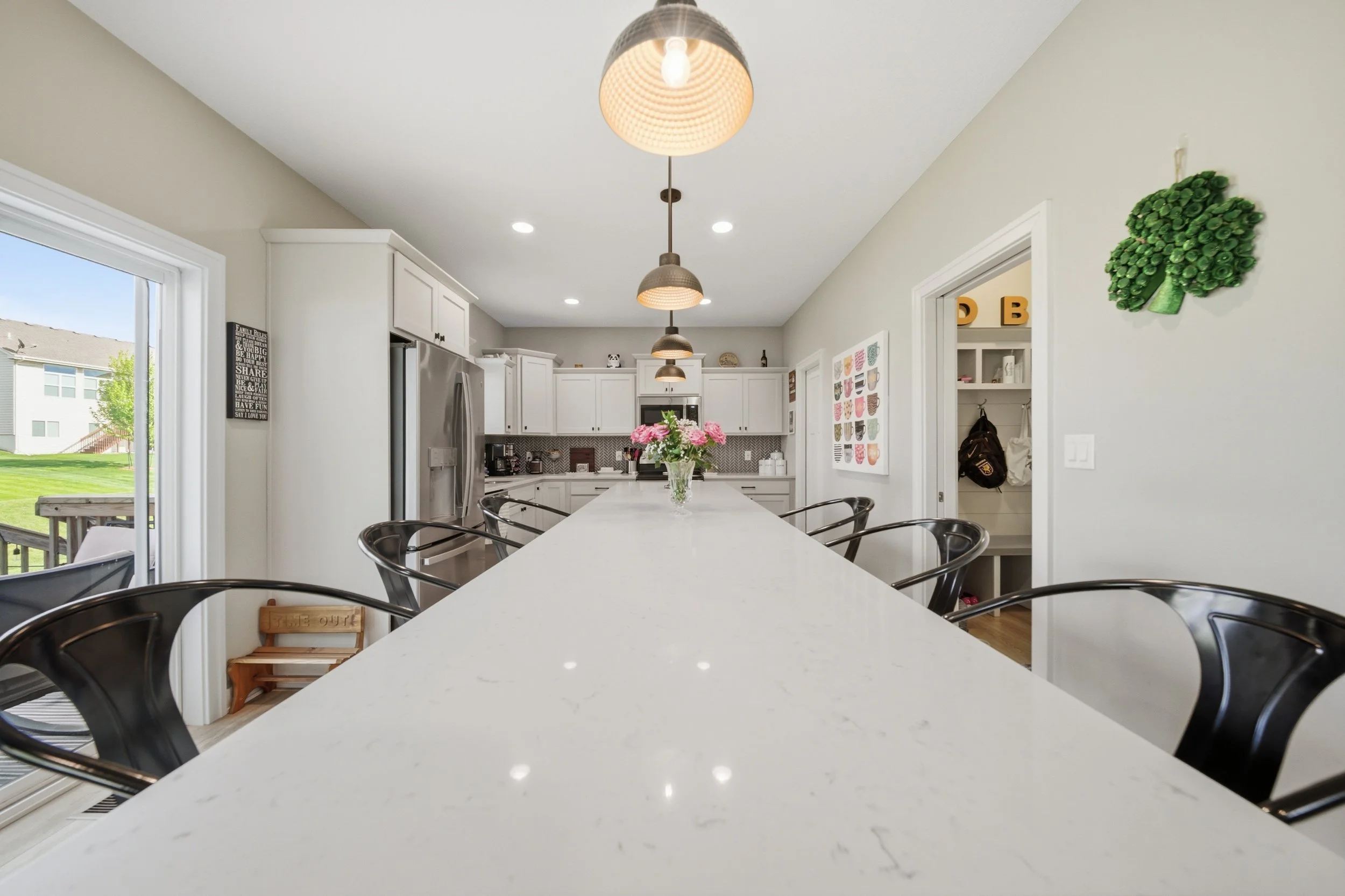 HDR real estate photography with a view of a bright, modern kitchen with white cabinets, pendant lights, a large white island with a vase of pink flowers, and barstools. Large window showing a backyard with grass and neighboring houses.