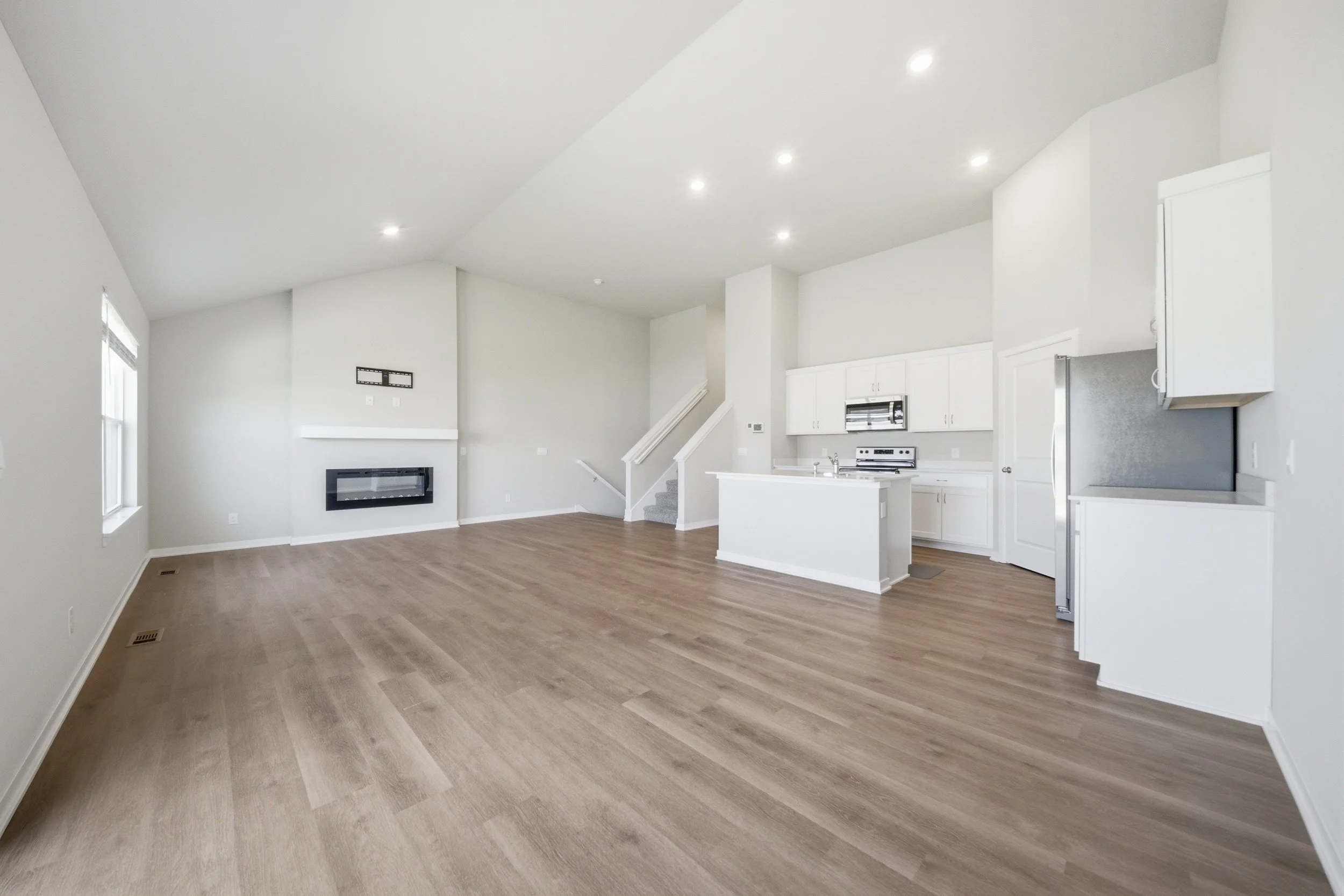 Real estate photography of an open-concept living area with light-colored walls, hardwood floors, a modern electric fireplace, and a kitchen with white cabinets and stainless steel appliances.