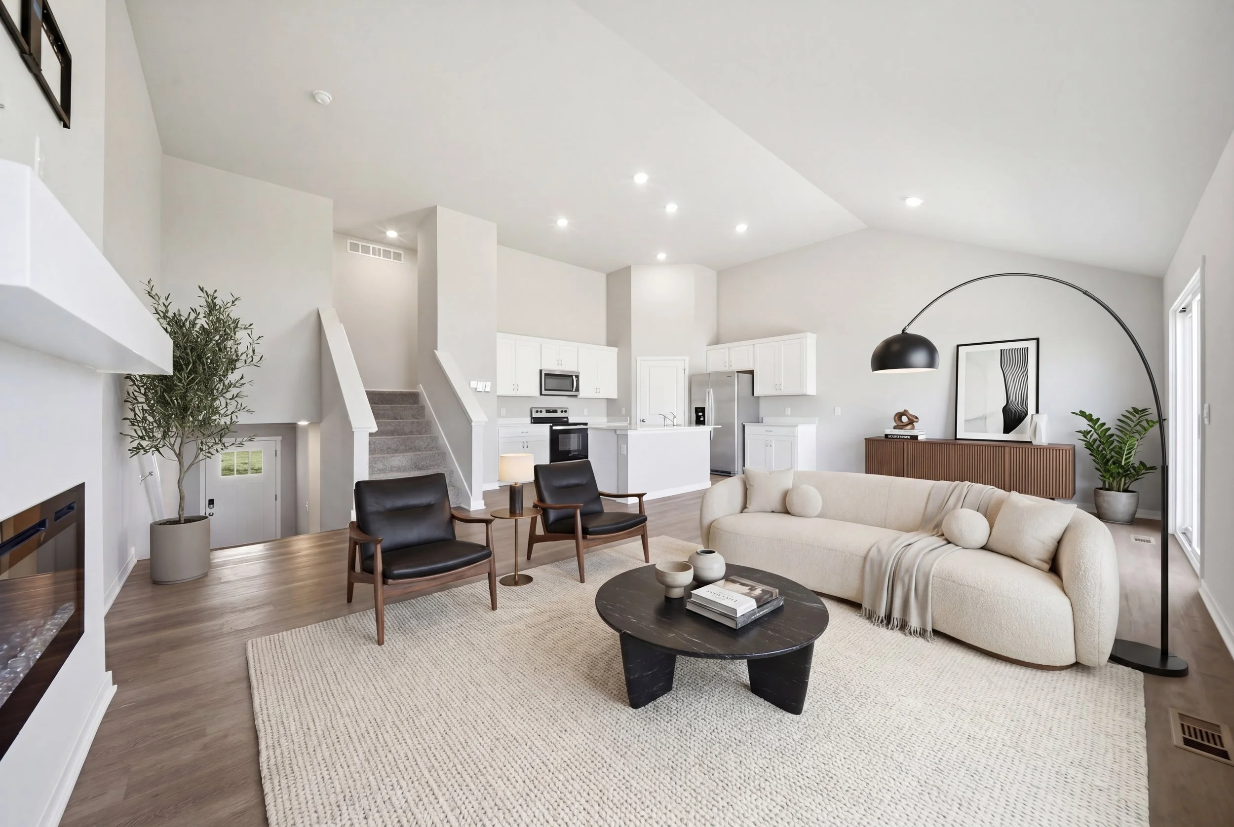 Real estate virtual staging photography of a living room with white walls, a beige sofa, black coffee table, and wooden chairs. Open kitchen with white cabinets, stainless steel appliances, and hardwood floors. 