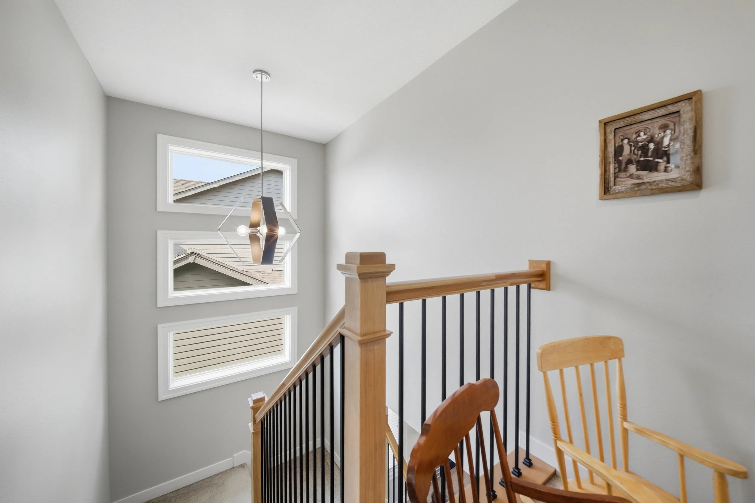 Interior view of a staircase with wooden handrail and black balusters, light gray walls, and three square windows on the wall. A modern geometric pendant light fixture hangs from the ceiling. Part of a wooden chair is visible in the foreground, and a