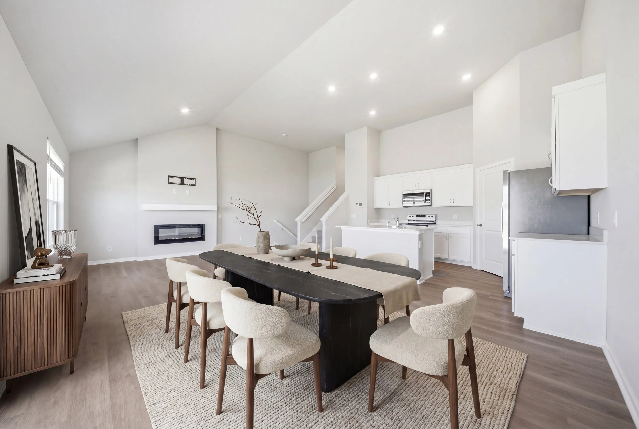 Real estate virtual staging photography of an open-concept kitchen and dining area with white walls, stainless steel appliances, a dining table with beige chairs, and a living space with a fireplace and artwork.