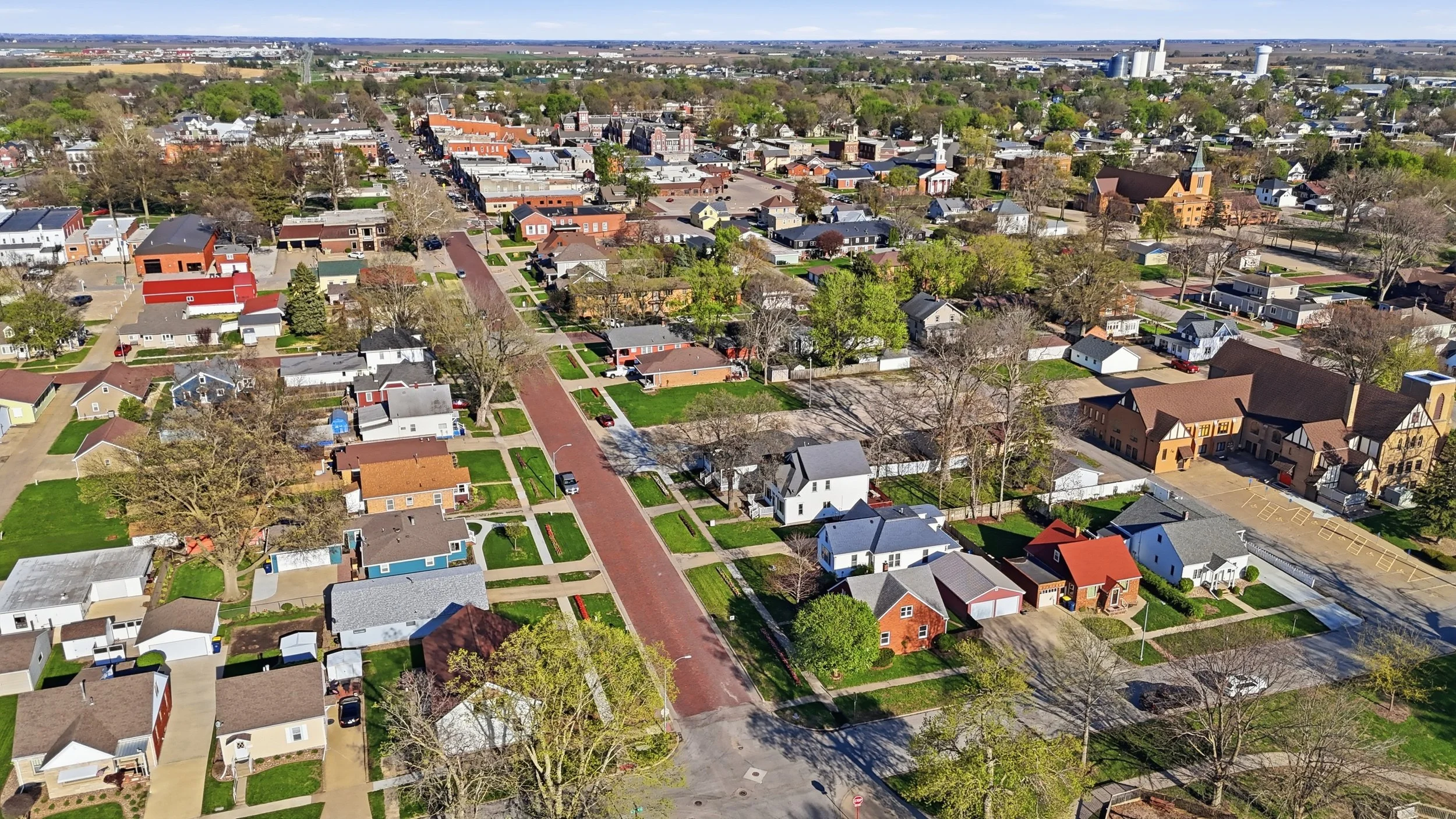 Real estate photography of an aerial view of a small town with residential houses, trees, and a few commercial buildings in the background, under a partly cloudy sky.