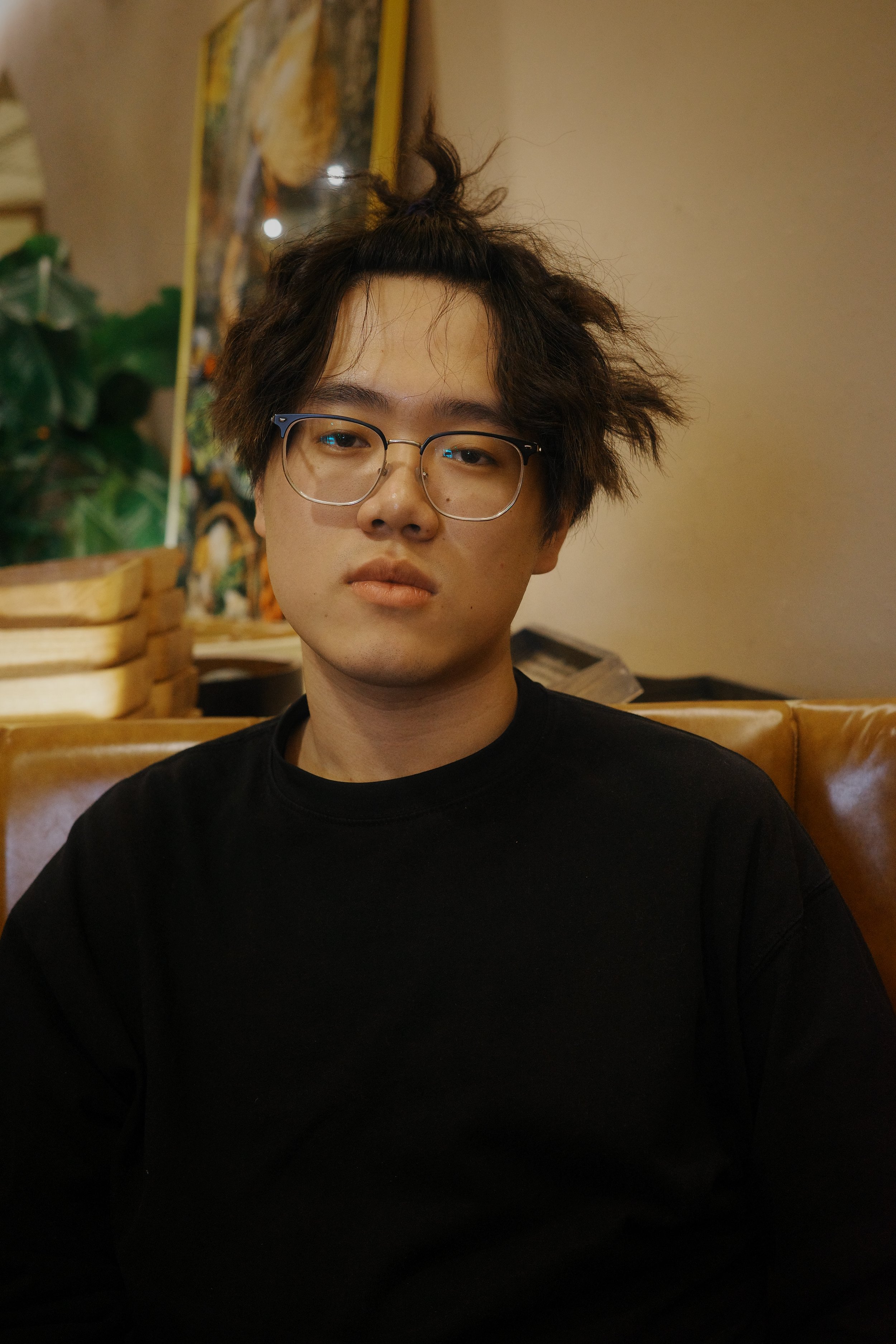 A young man with tousled dark hair, wearing glasses and a black shirt, sits on a brown leather couch inside a cafe or restaurant with a colorful painting and green plant in the background.