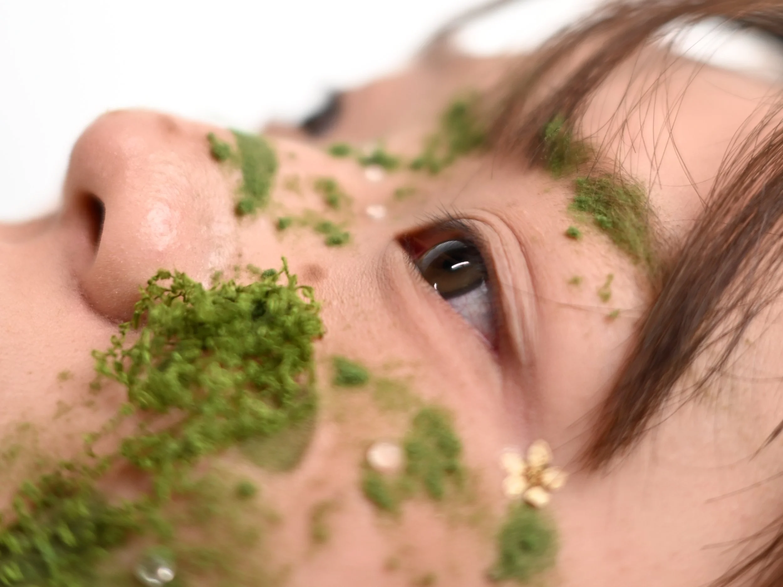 Close-up of a woman's face with green moss on her nose, forehead, and cheek, lying down with her eye partially open.