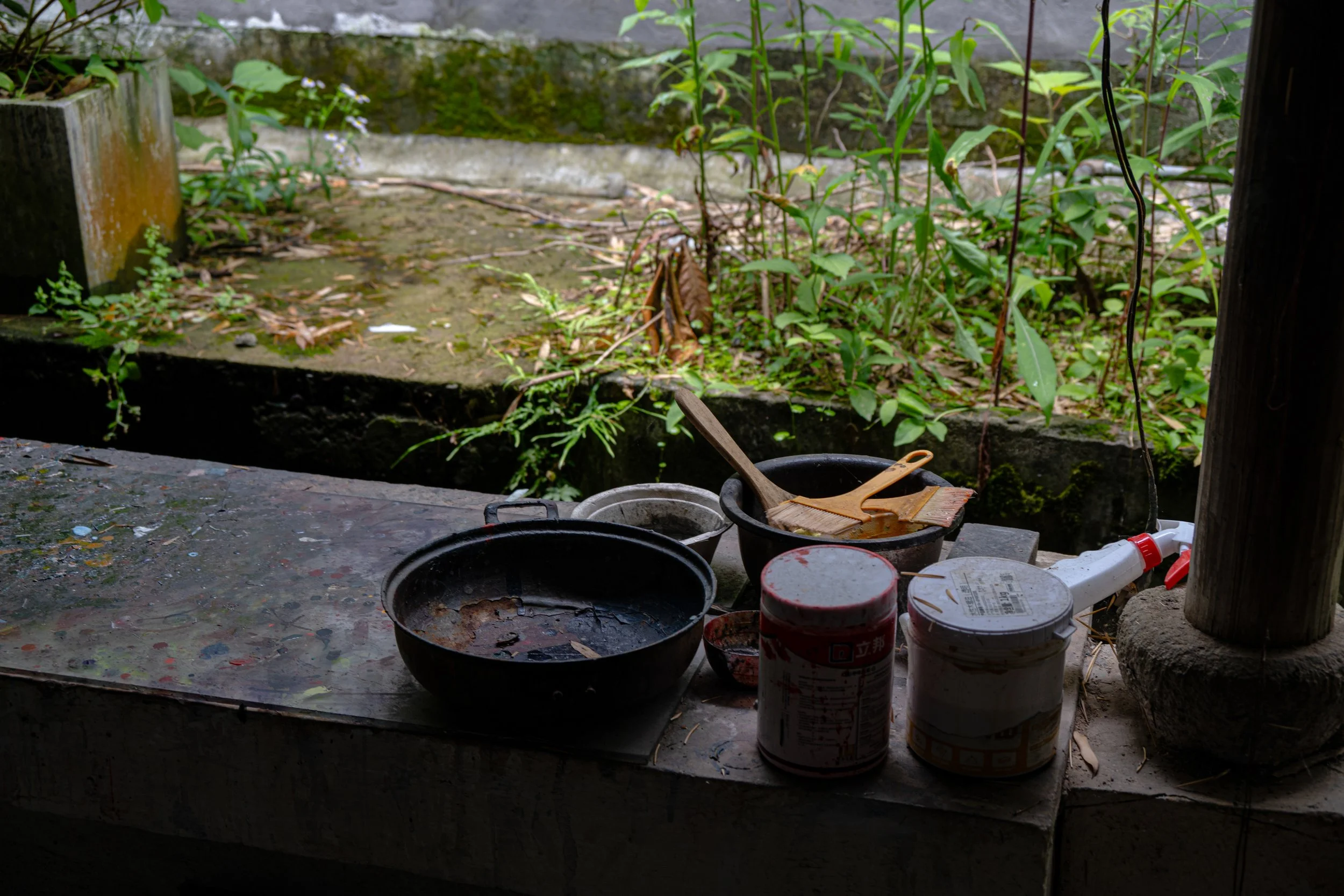 Outdoor kitchen area with empty black cooking pans, wooden utensils, and containers on a weathered stone countertop, overlooking a garden with green plants.
