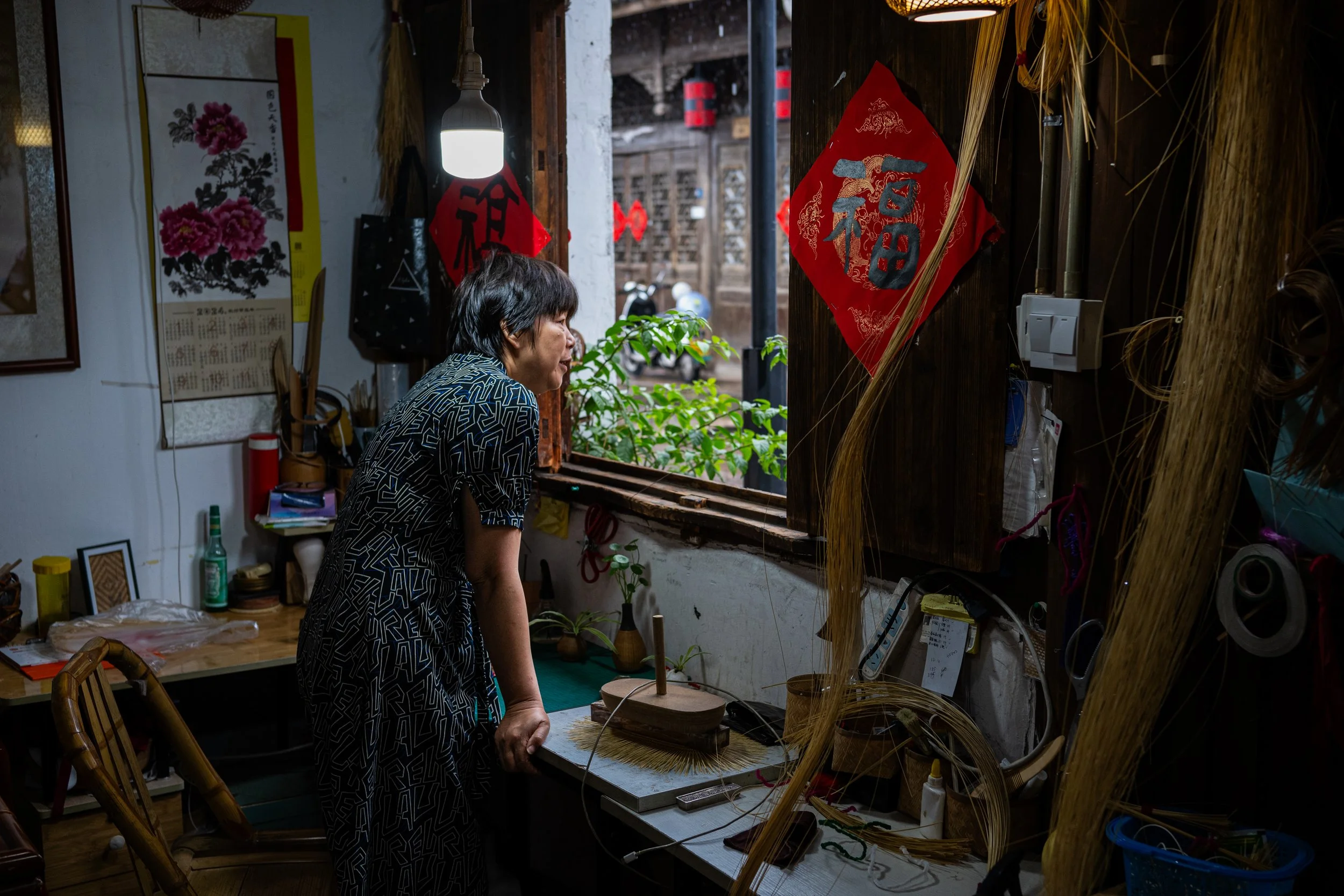 A woman in a patterned dress leaning over a table inside a room decorated with red Chinese New Year decorations, looking out a window at the street outside.