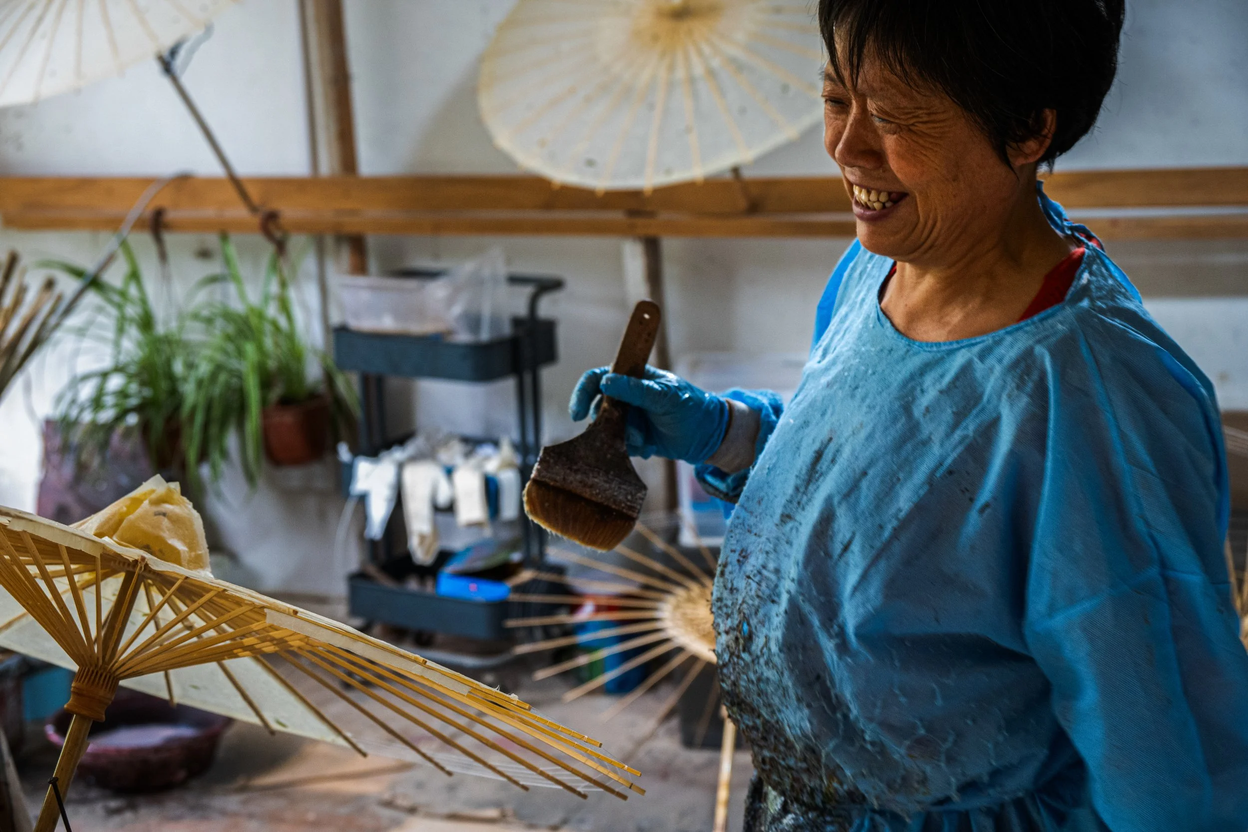 A smiling woman in a blue apron and gloves holding a brush, working with paper umbrellas in a workshop.