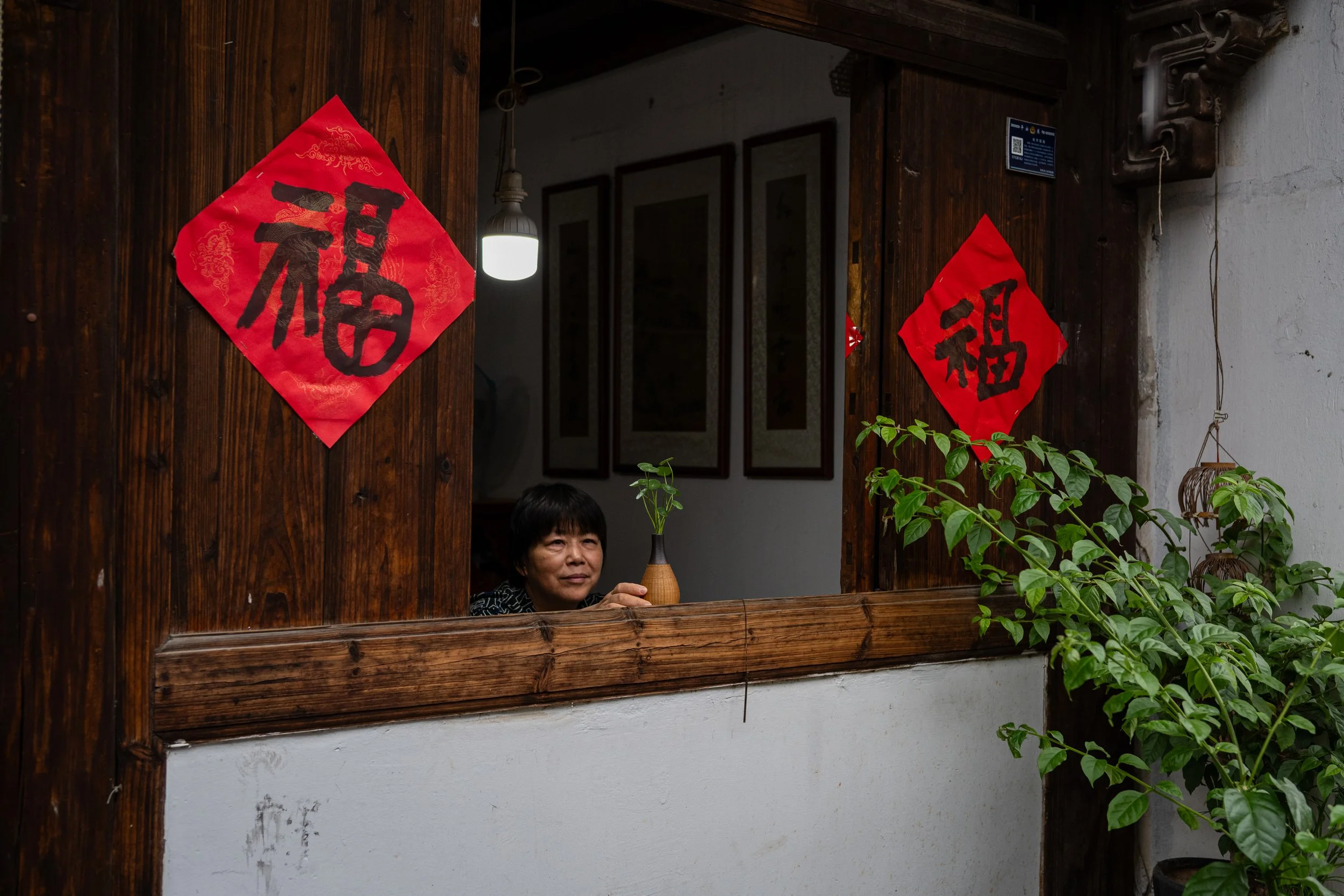A woman peeking through a window with two red Chinese New Year decorations featuring the character '福' (fortune) on either side. There are plants and framed artworks in the background.