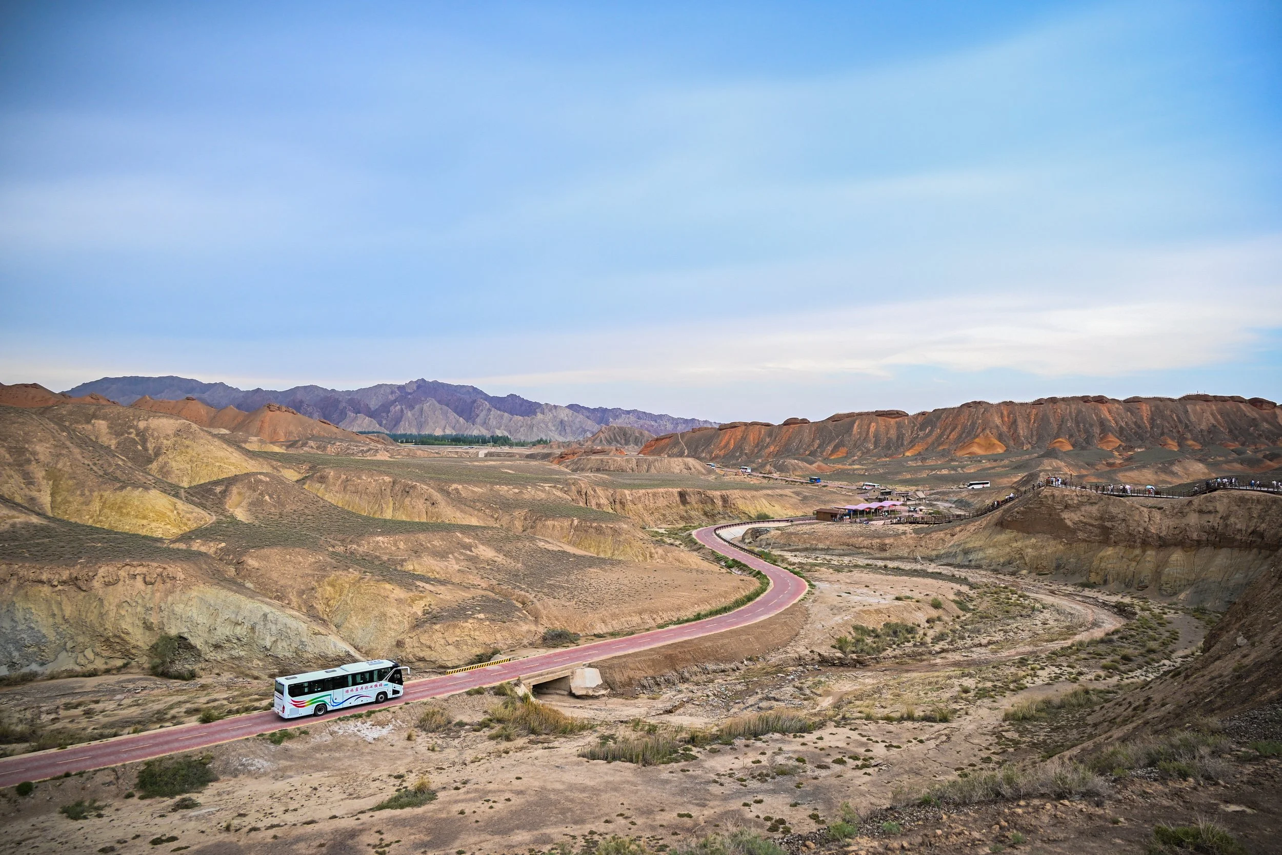 A bus traveling on a winding road through arid, colorful desert hills with mountains in the distance under a blue sky.