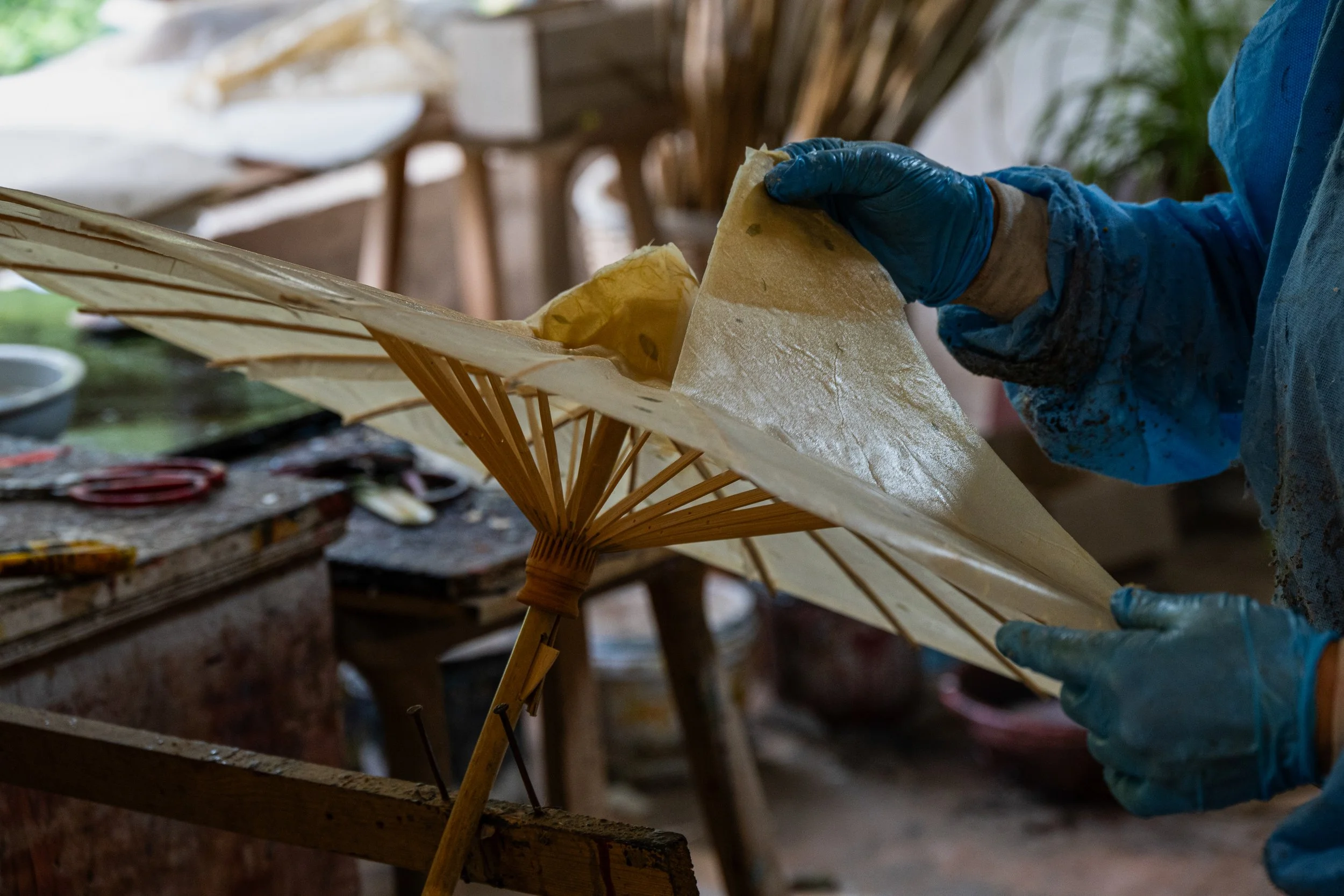 A person wearing blue gloves is working on a traditional Asian paper parasol, applying a glossy finish to the paper canopy in a workshop.