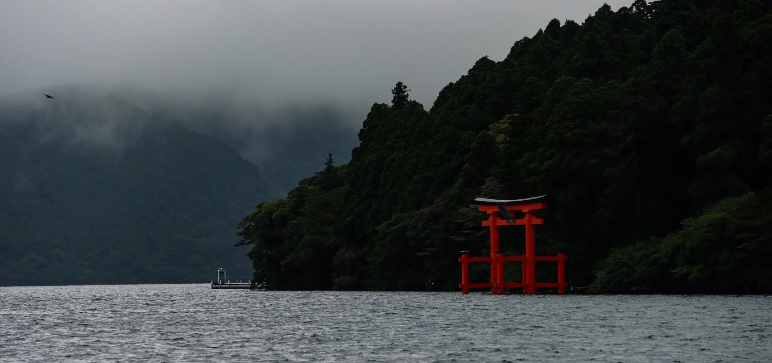 A red torii gate partially submerged in water near a forested hillside, with misty mountains and a cloudy sky in the background.