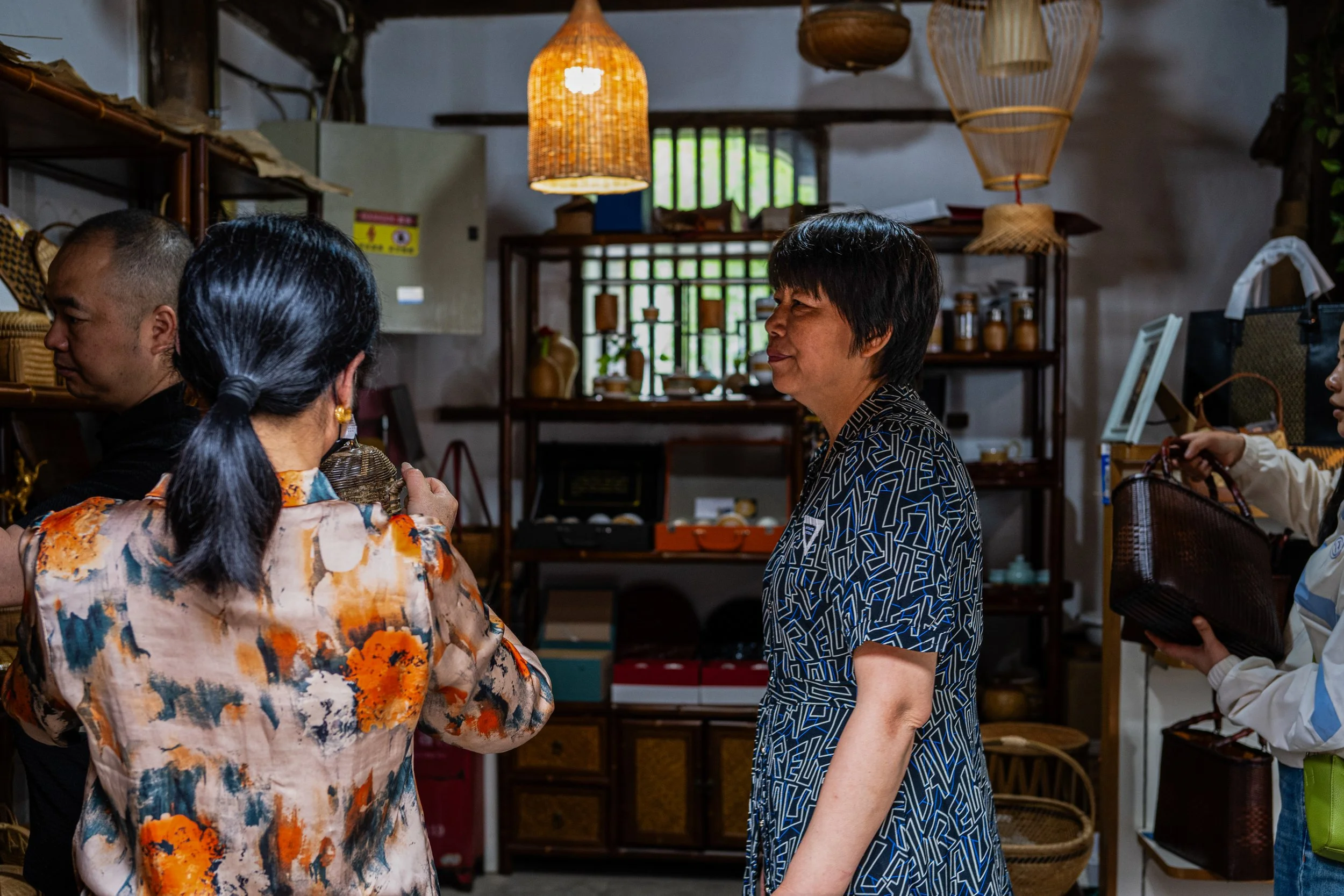 A group of four women in a shop, with one woman facing the others, engaging in conversation, inside a store with wooden shelves and hanging baskets.