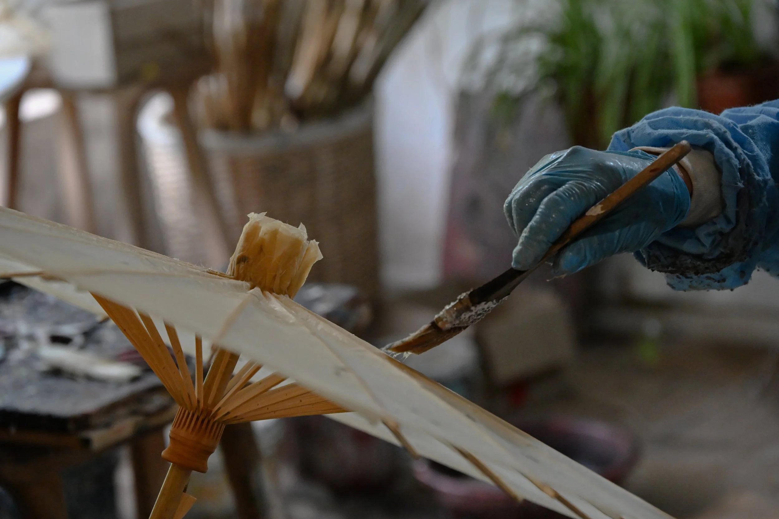 A person wearing blue gloves and a long-sleeve shirt is holding a paintbrush and working on a parasol or umbrella, likely painting or applying a coating. In the background, there are wicker chairs and a green potted plant.