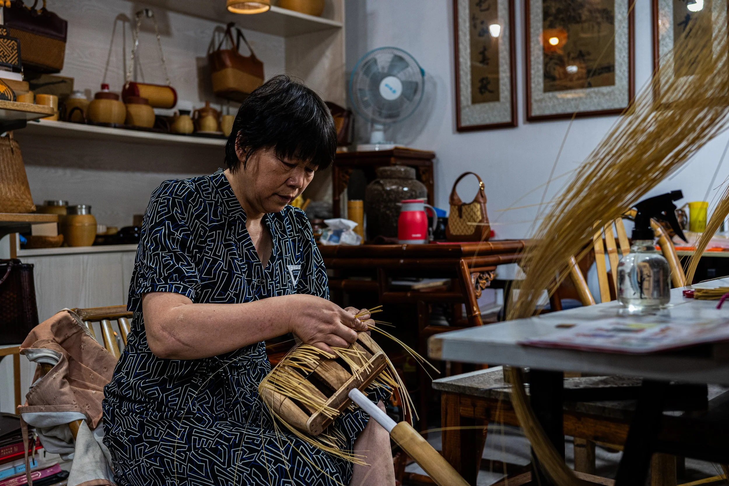 A woman weaving with straw in a room filled with woven baskets and traditional items.